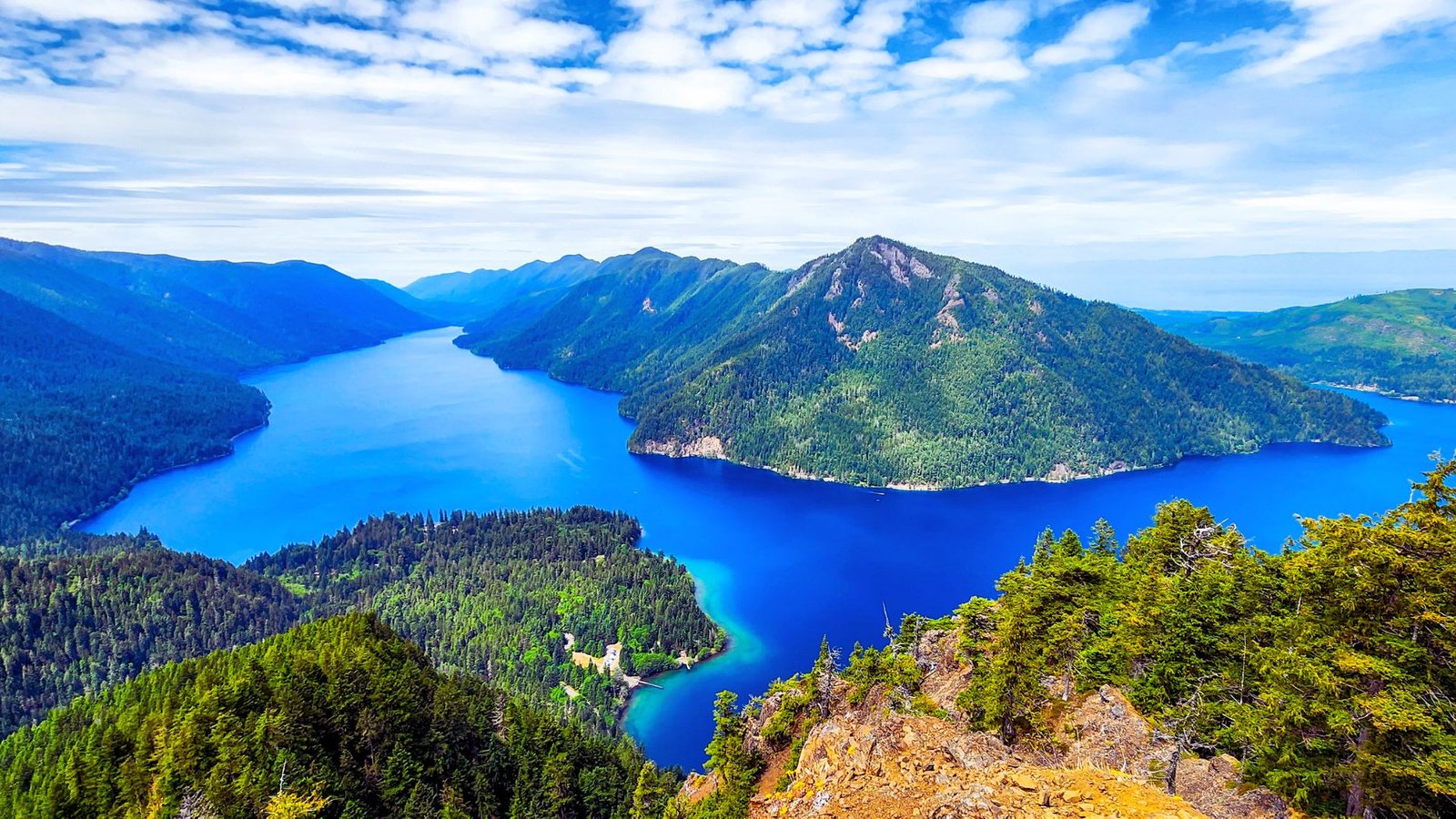 A panoramic view from a mountain peak, showcasing a serene blue lake surrounded by lush greenery.