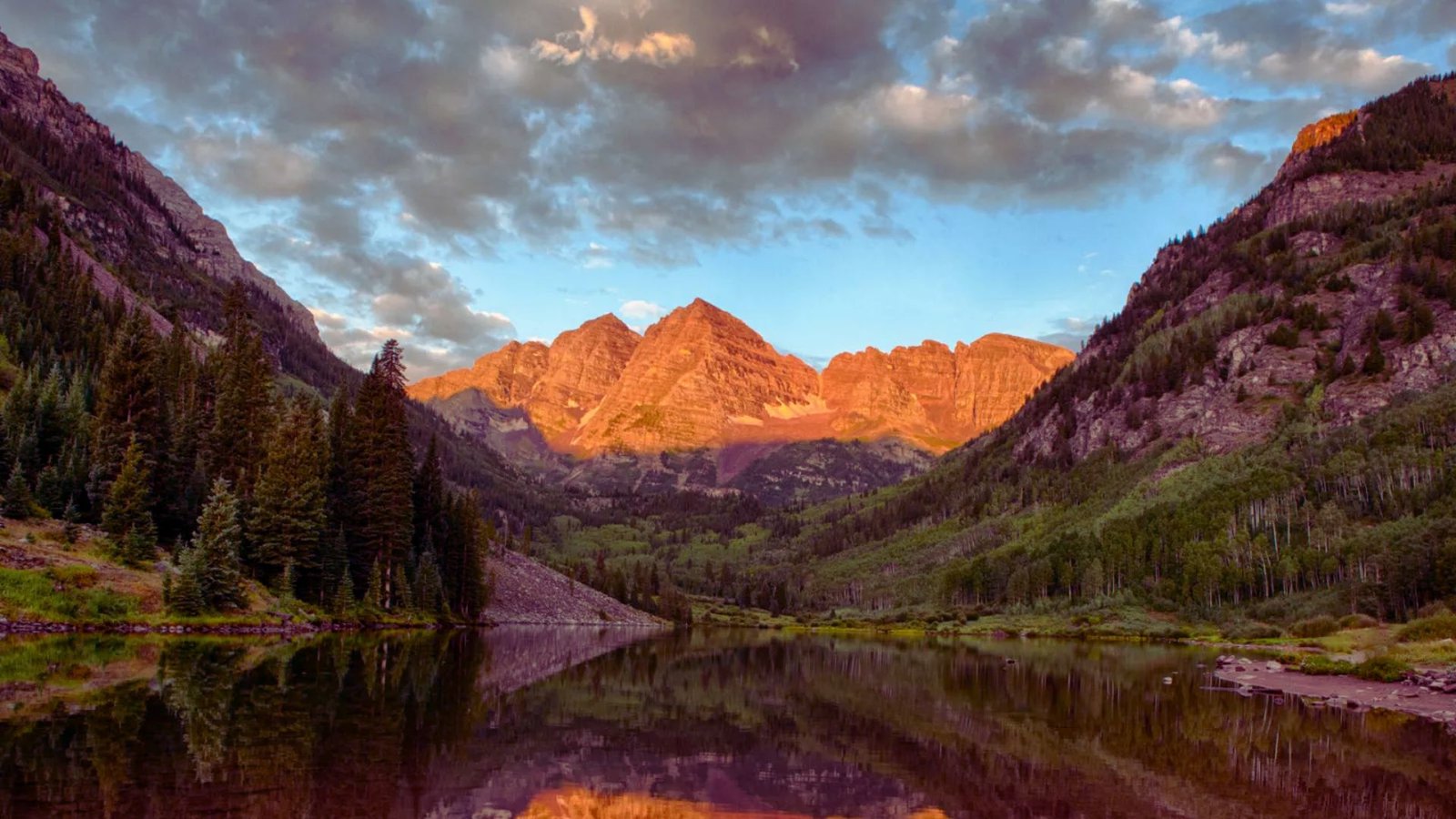The maroon bells reflecting in a tranquil lake at sunset, surrounded by vibrant autumn foliage and a colorful sky.