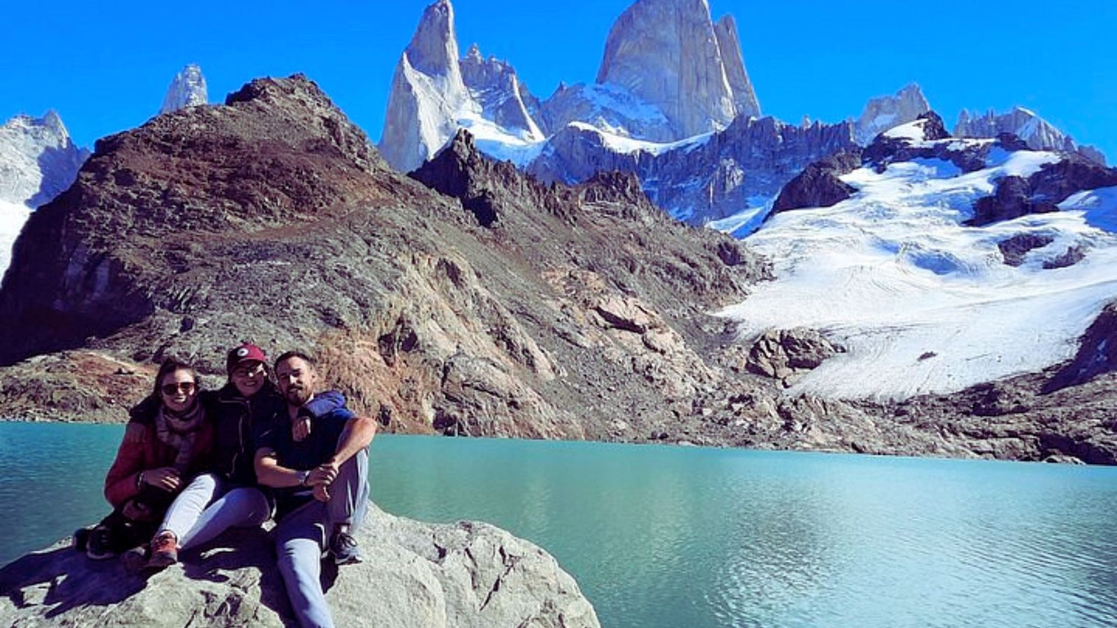  Three people sitting on a large rock by a serene lake, enjoying the view and each other's company.
