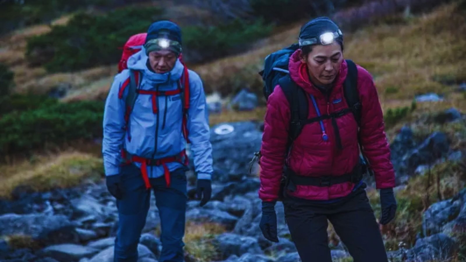 Two individuals with headlamps navigate a rocky trail under low light conditions.