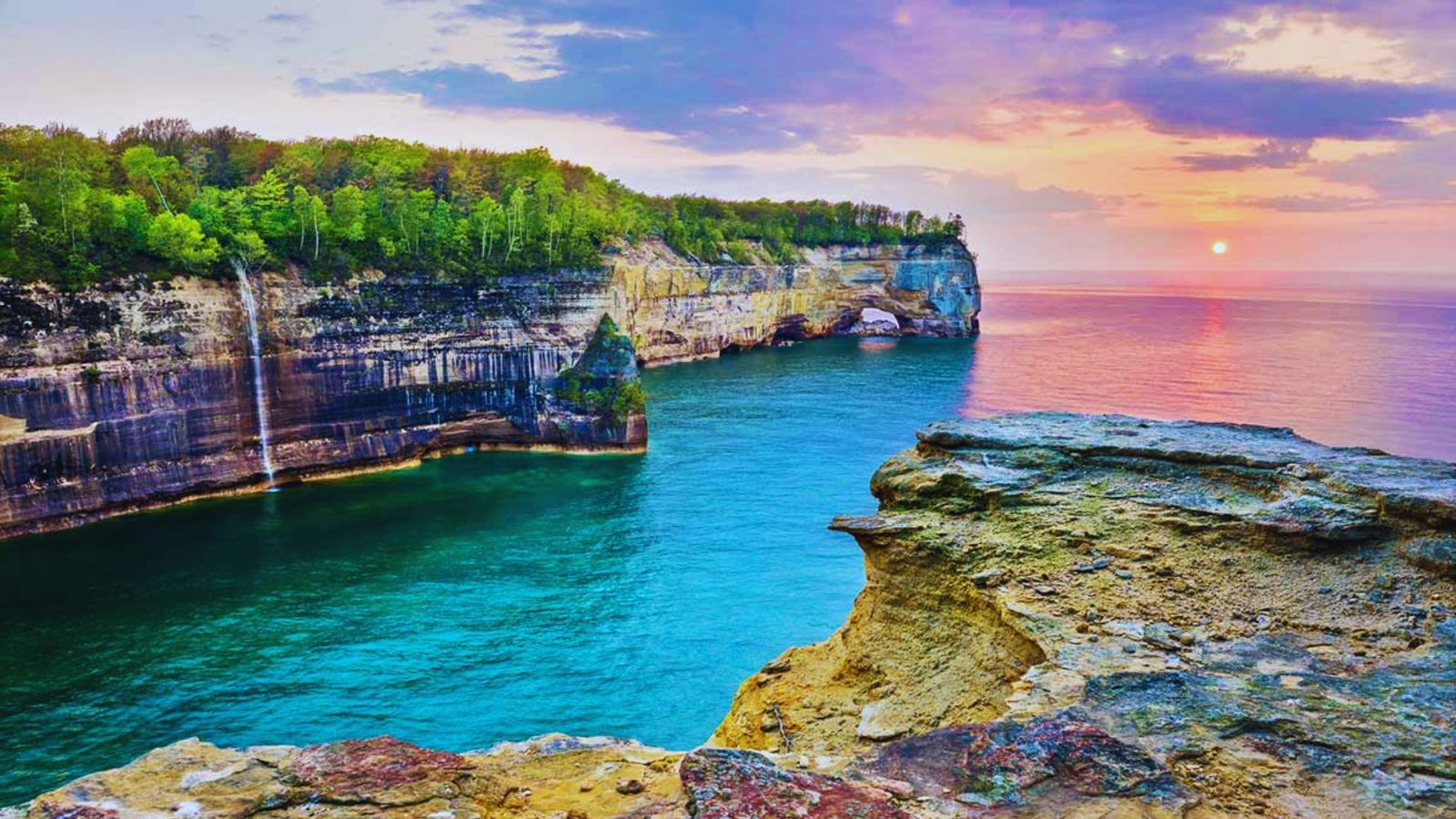 The sun sets over the cliffs at Pictured Rocks National Lakeshore, casting vibrant colors across the sky and water.