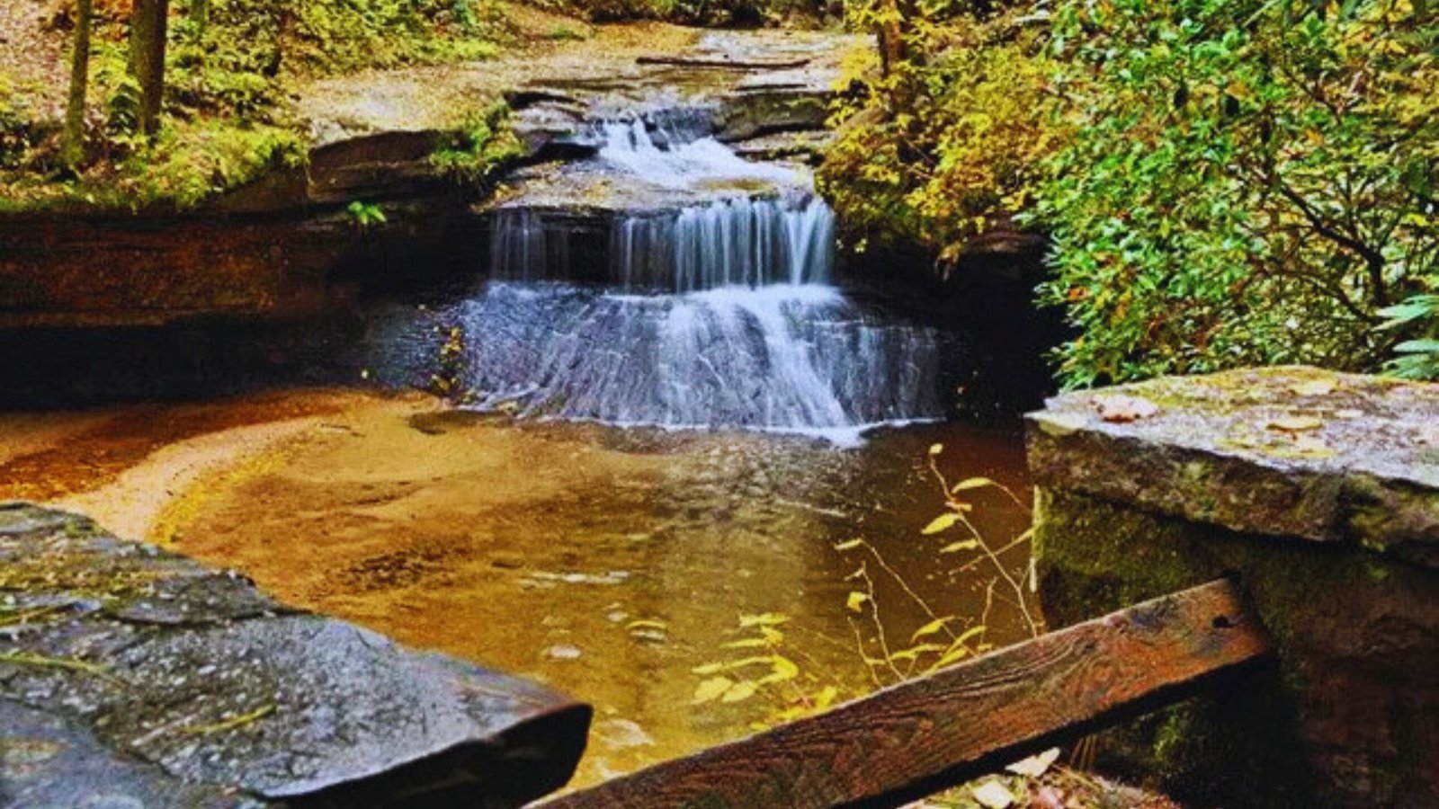 A serene waterfall cascades through the woods, with a rustic wooden bridge crossing over the flowing water.