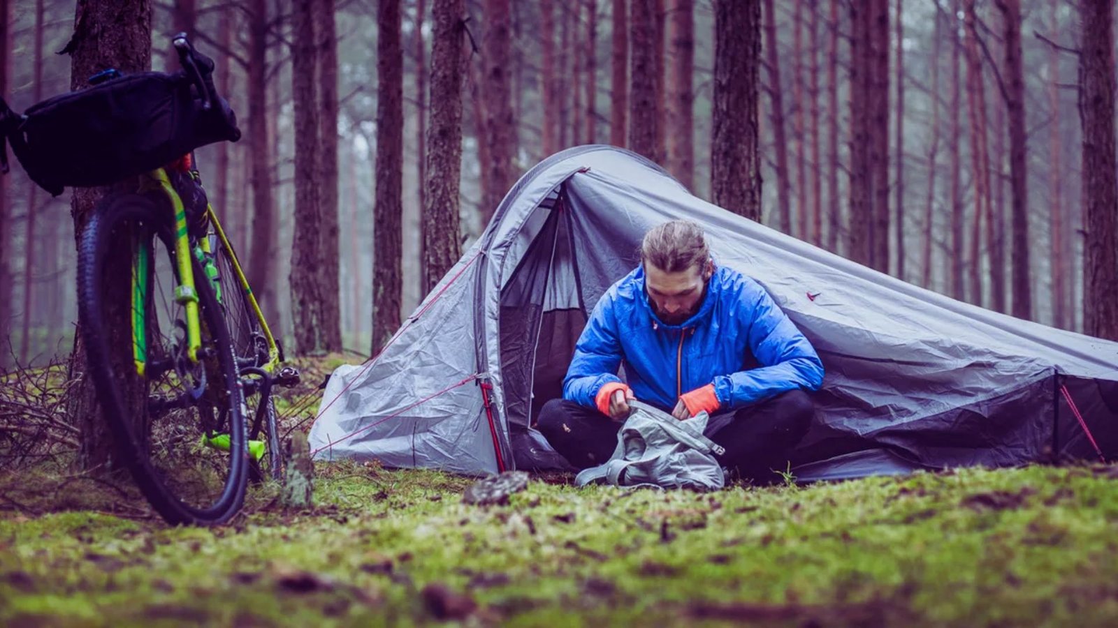A man is assembling a tent in a wooded area, surrounded by trees and natural scenery.
