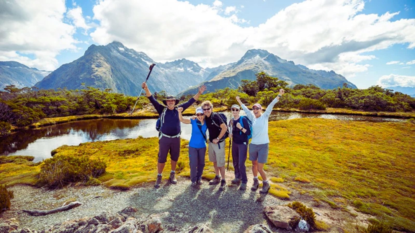 Four people smile for a photo in front of a serene mountain lake, surrounded by lush greenery and distant peaks.
