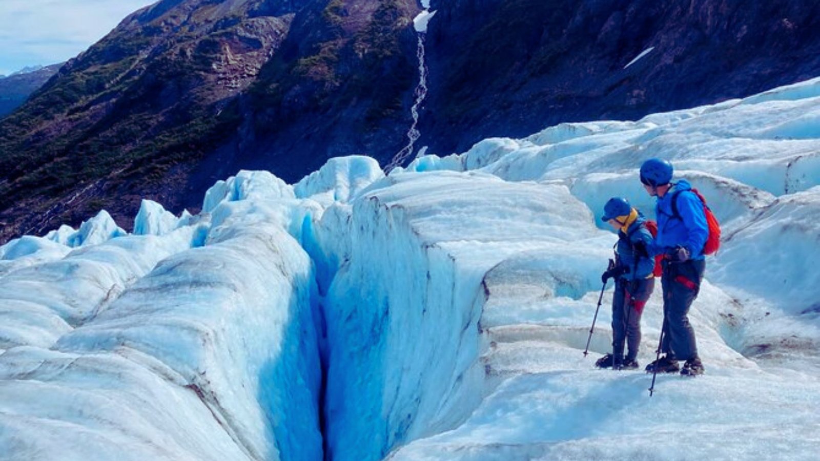 Two hikers navigate a glacier, surrounded by icy terrain and snow-capped peaks under a clear blue sky.