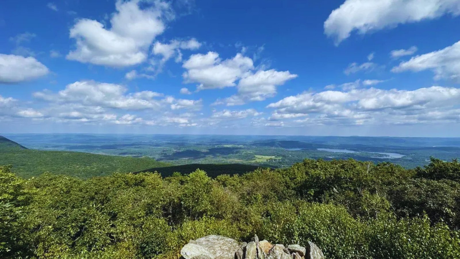A panoramic view from a mountain peak, showcasing a serene lake surrounded by lush greenery and distant hills.
