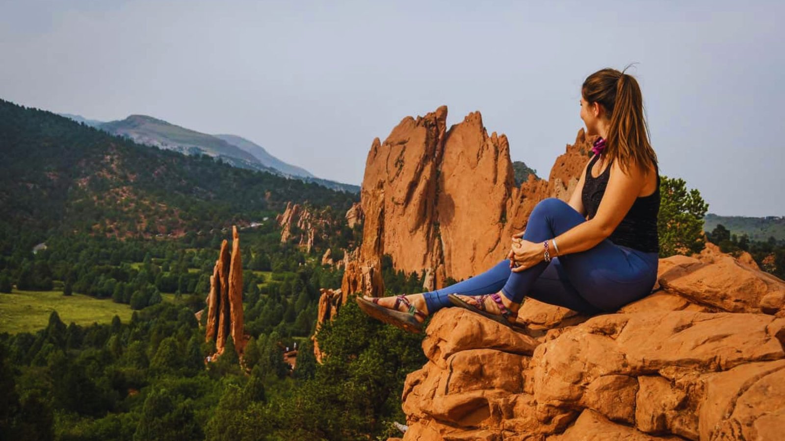 A woman sits on a rock, gazing at the expansive mountains in the distance under a clear blue sky.