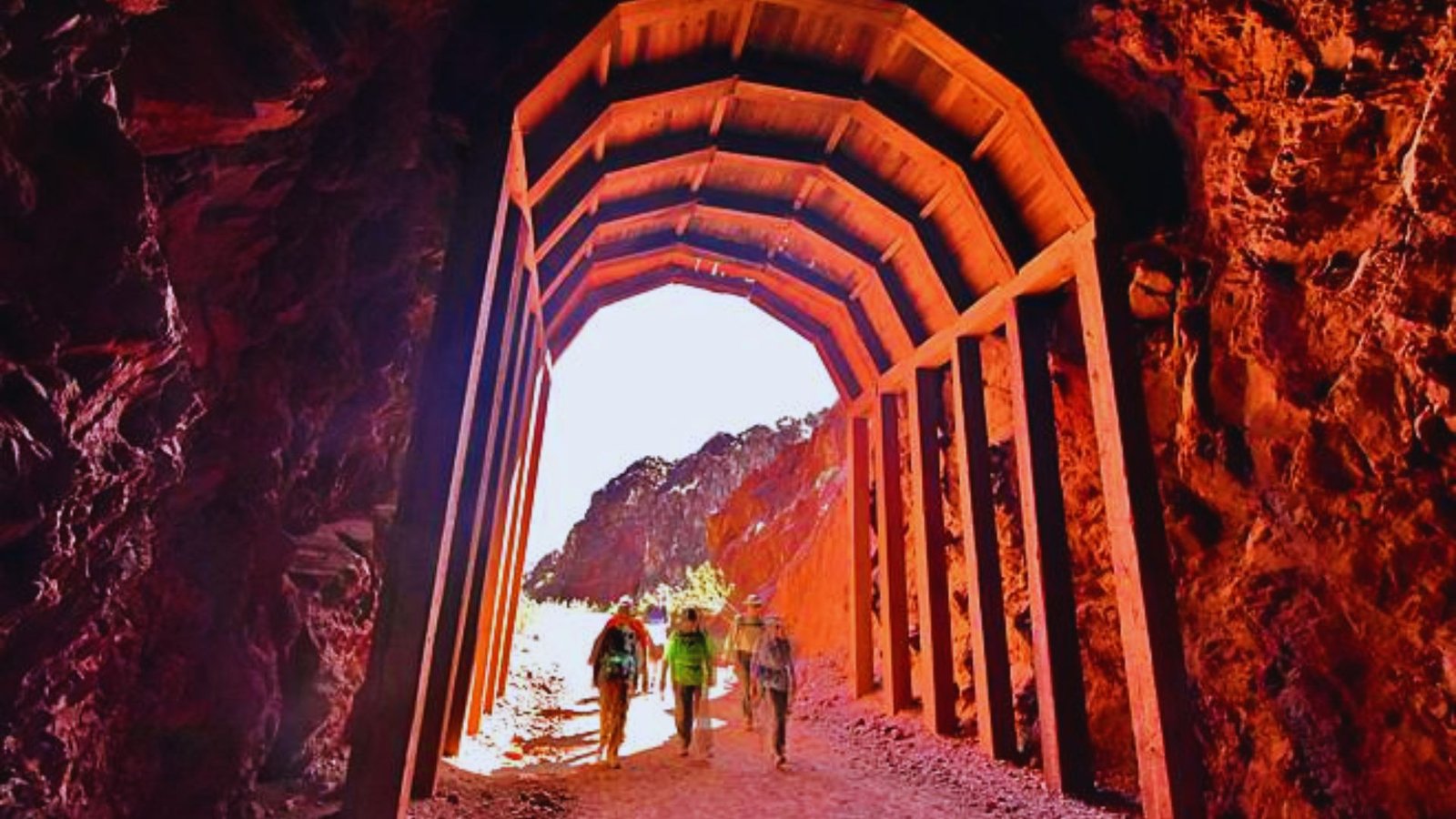 Three people walk through a stone archway in a vast desert landscape under a clear blue sky.
