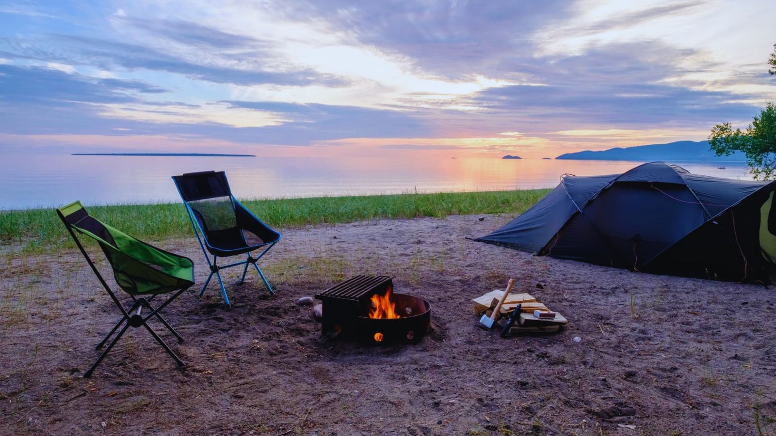 A tent and chair are set up on a beach, with a vibrant sunset casting warm colors over the serene scene.
