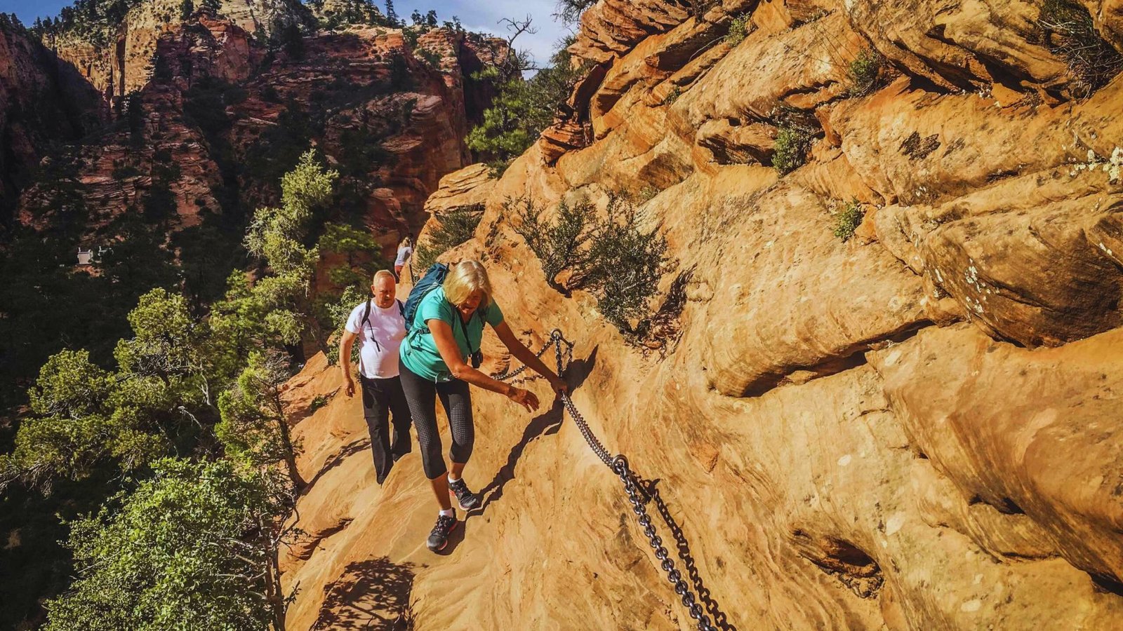 Two individuals trekking along a narrow trail in the mountains, with steep slopes and scenic views.