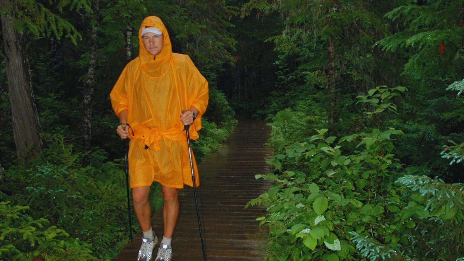 A man in an orange raincoat walks along a wooden path surrounded by greenery.