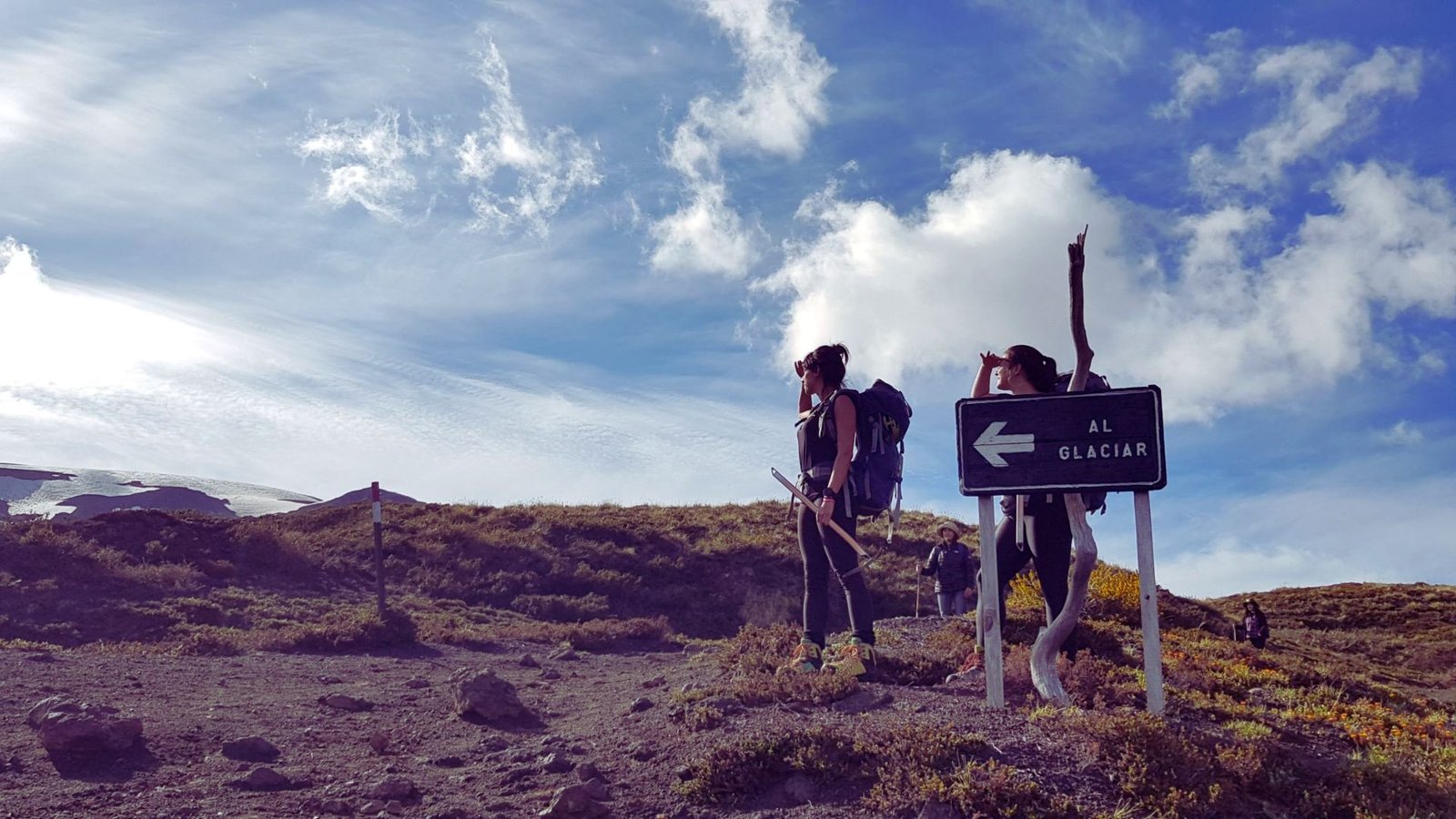 Two people stand beside a mountain sign, surrounded by rocky terrain and a clear blue sky in the background.