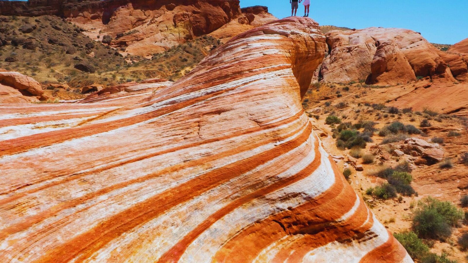 Two people stand atop a rock formation in Valley of Fire State Park, surrounded by vibrant red sandstone cliffs.
