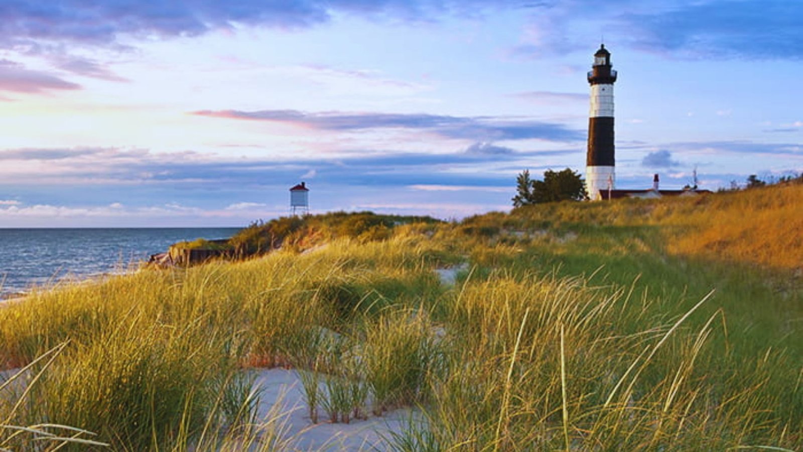 A lighthouse stands on a beach, surrounded by tall grass, with waves gently lapping at the shore in the background.