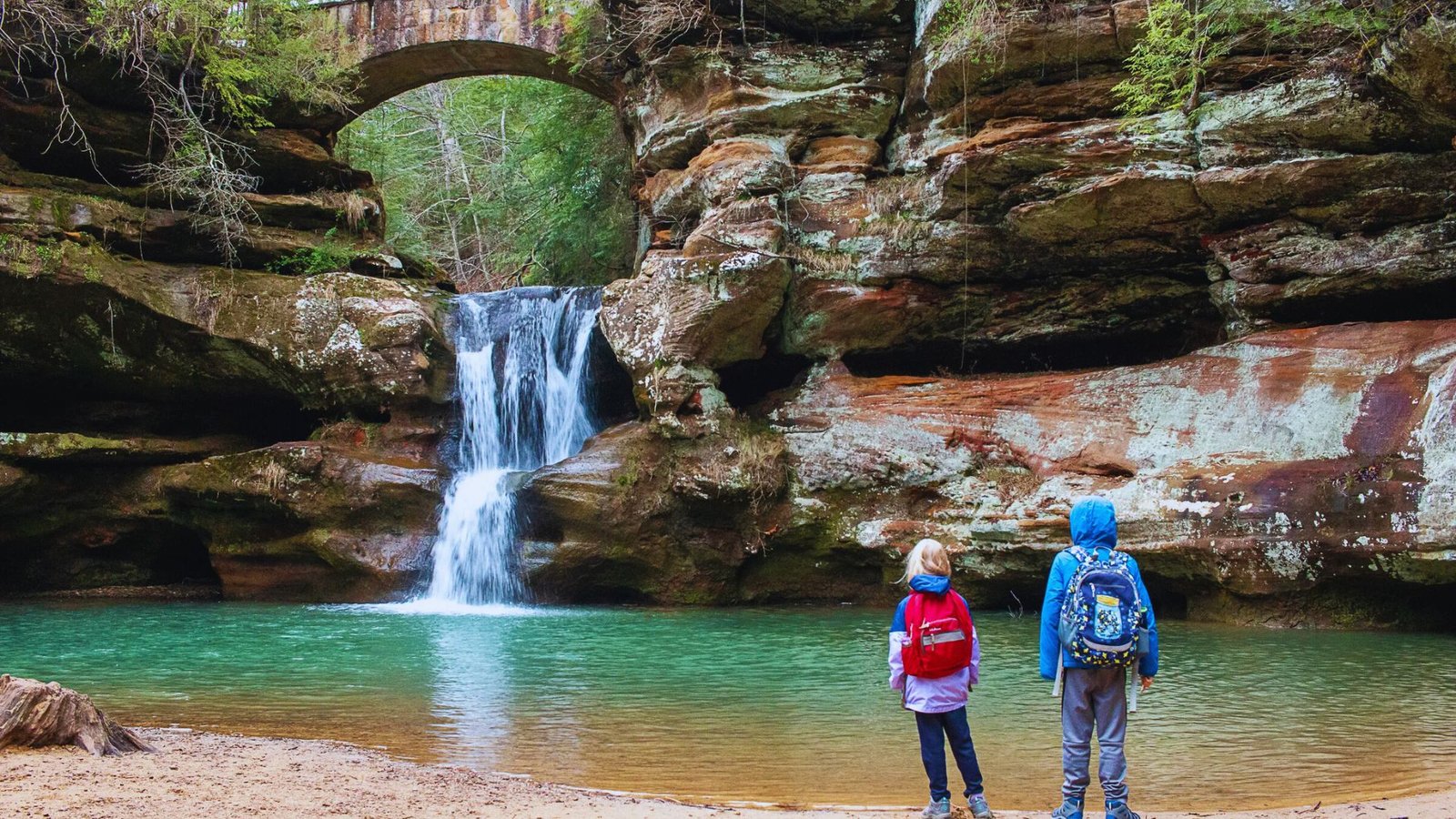Two children stand smiling in front of a cascading waterfall, surrounded by lush greenery.