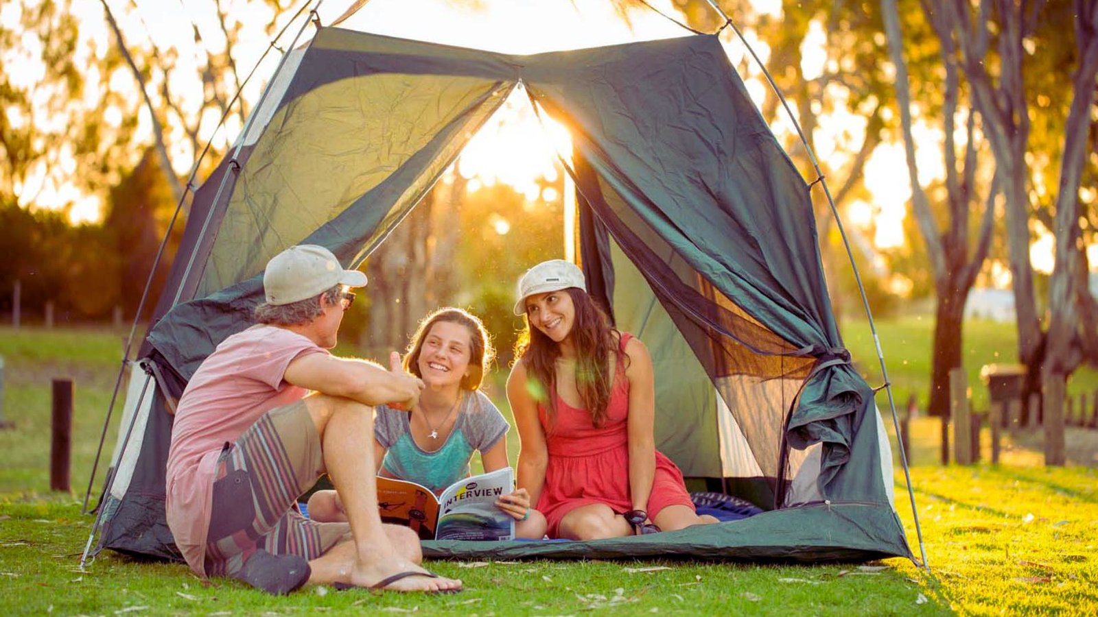 Three people sitting inside a tent, reading a book together, surrounded by camping gear and a cozy atmosphere.
