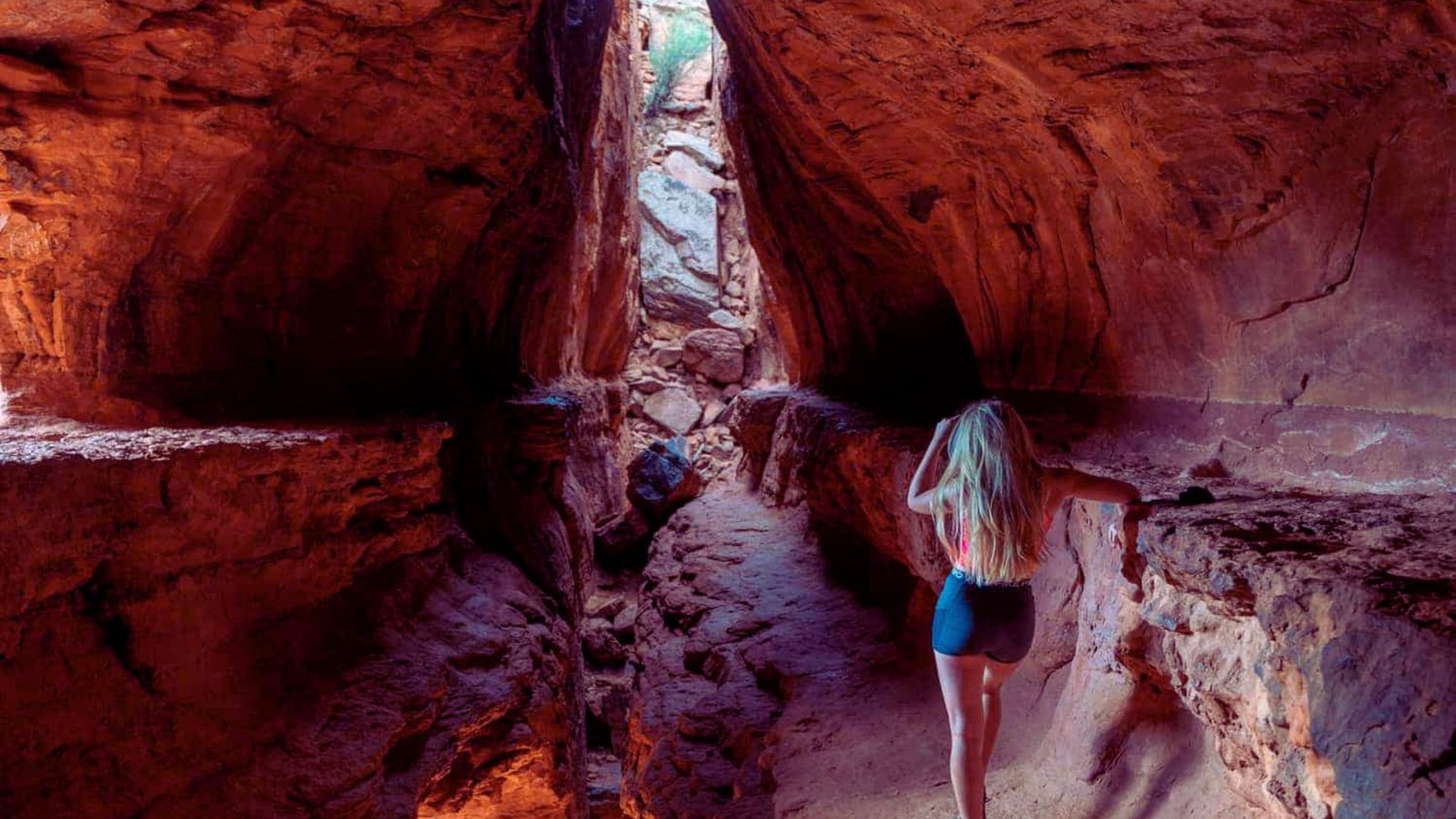 A woman walks through a narrow canyon, surrounded by towering rock walls and natural sunlight filtering down.