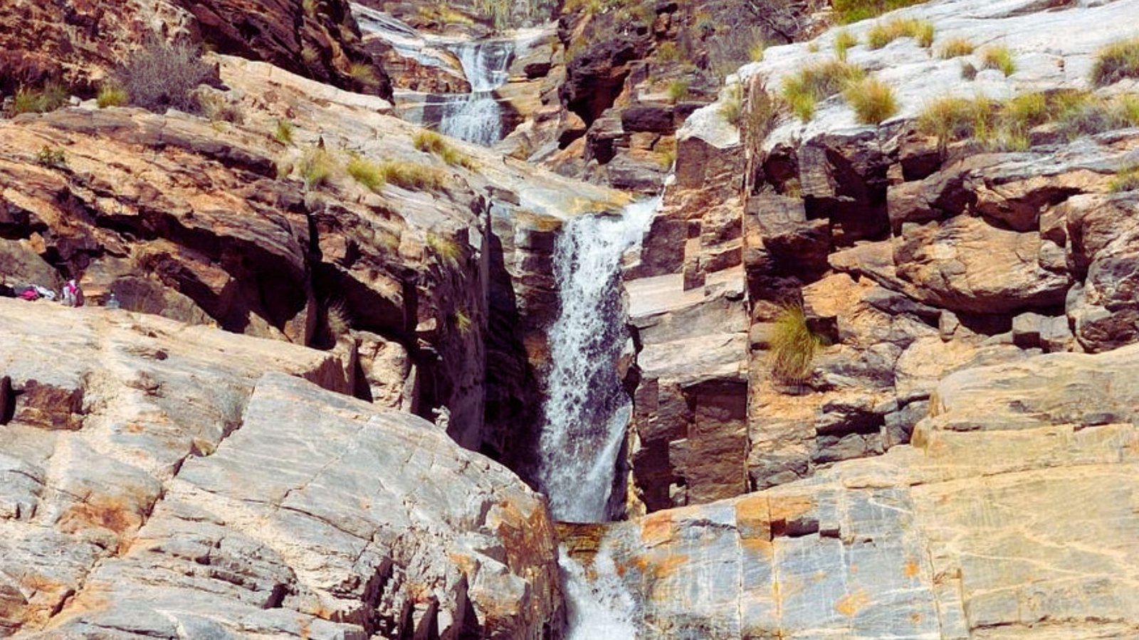 A waterfall cascades down a rugged mountain, surrounded by rocky terrain and lush greenery.