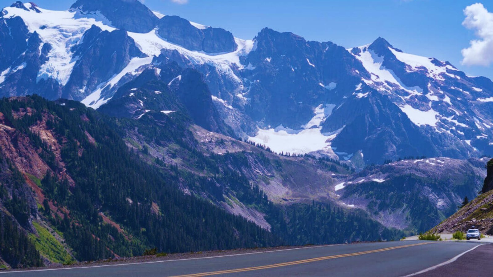 A car drives along a winding mountain road with snow-capped peaks visible in the background.