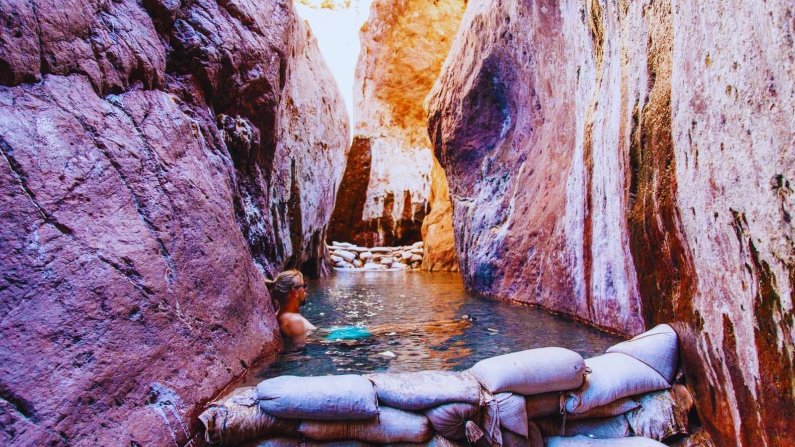 A man swims in a narrow canyon, surrounded by steep rock walls and clear blue water.
