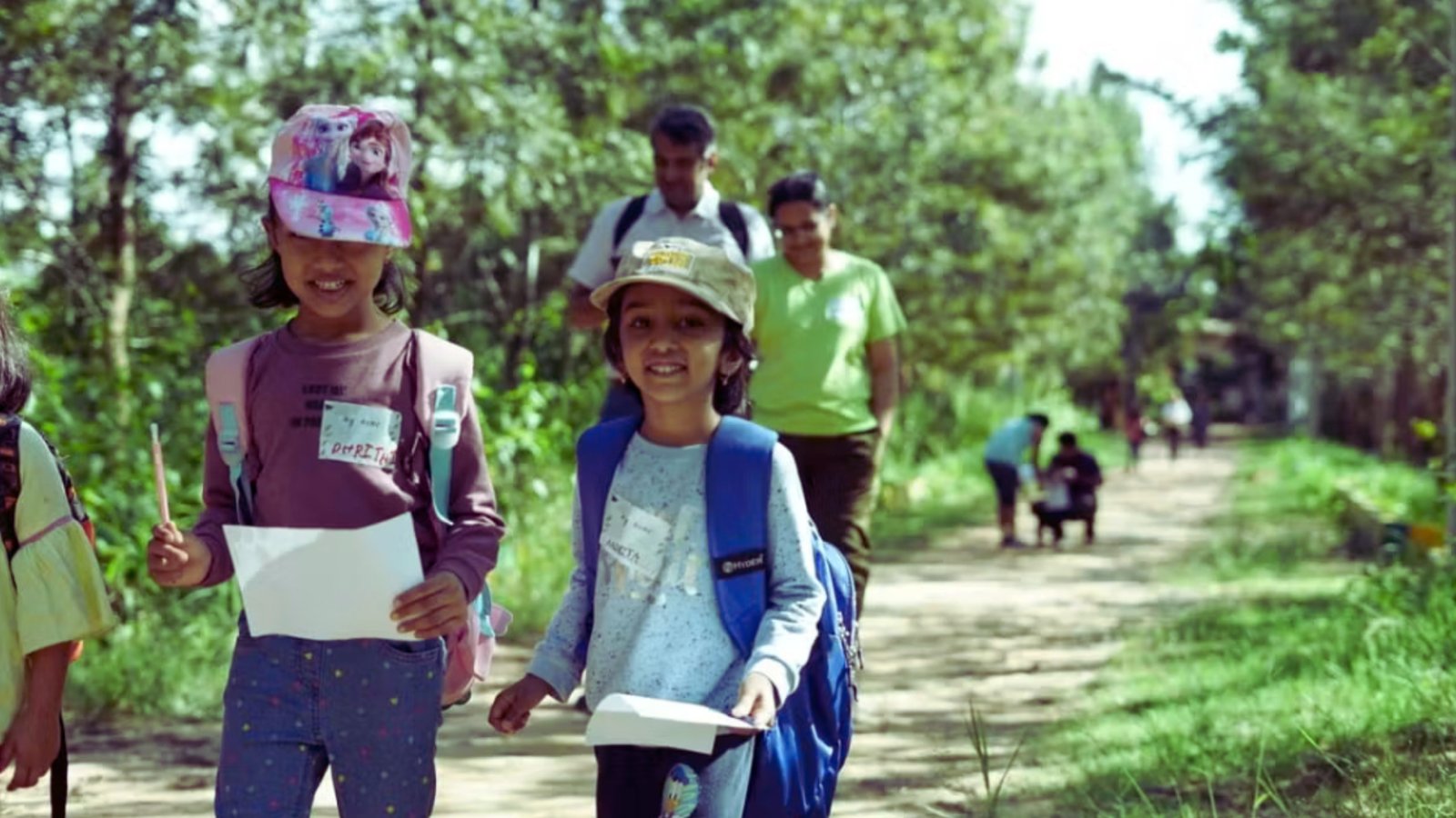  A group of children walking together along a dirt road, surrounded by greenery and a clear blue sky.