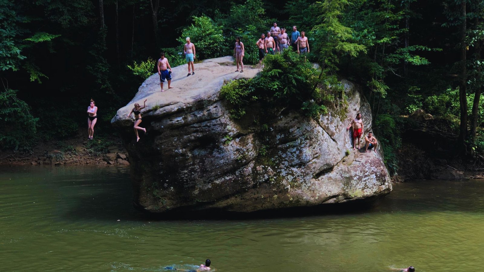 A group of people swimming in clear water near a large rock formation under a bright blue sky.