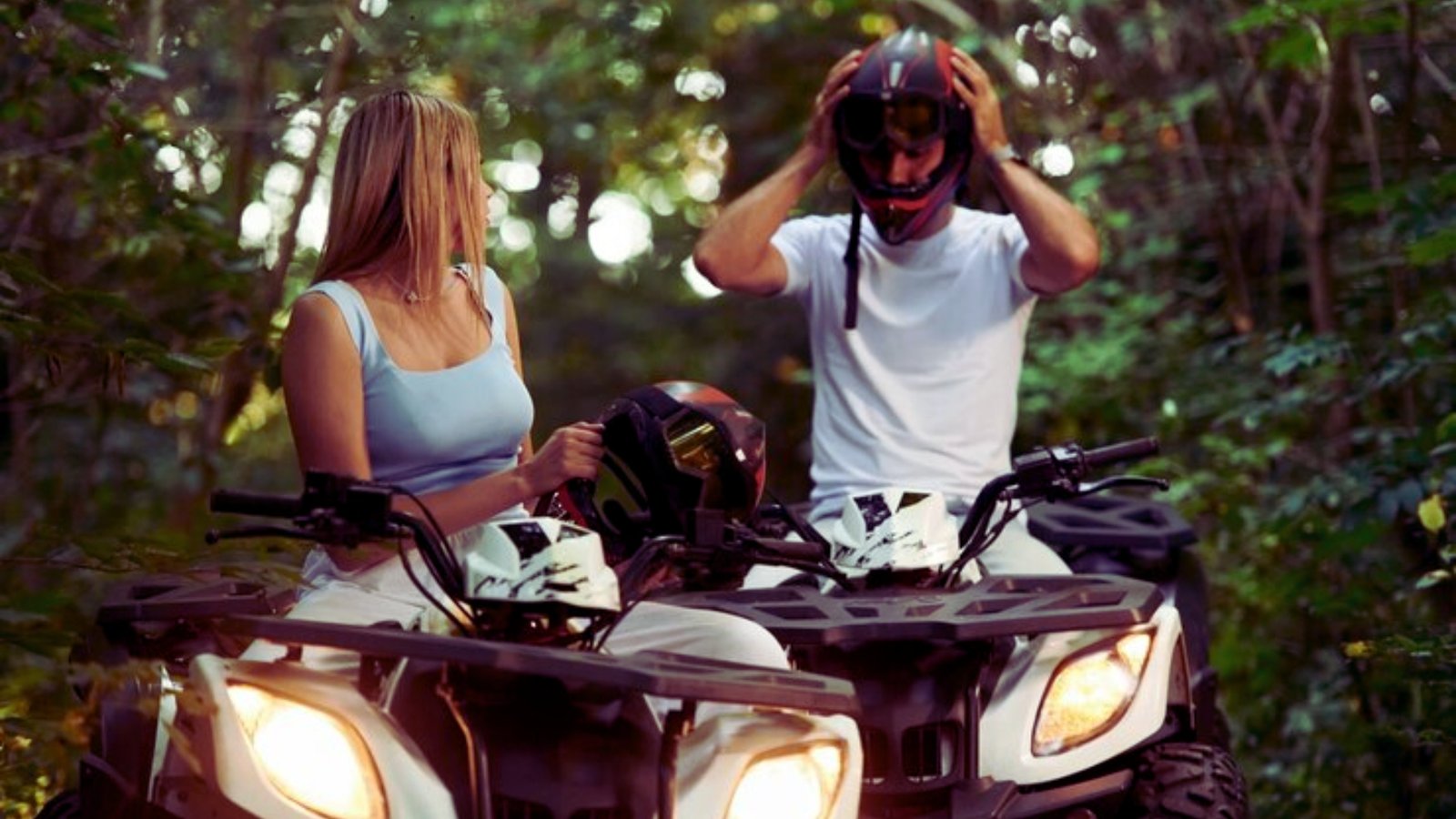 A man and woman riding all-terrain vehicles through a dense forest, surrounded by trees and natural scenery.