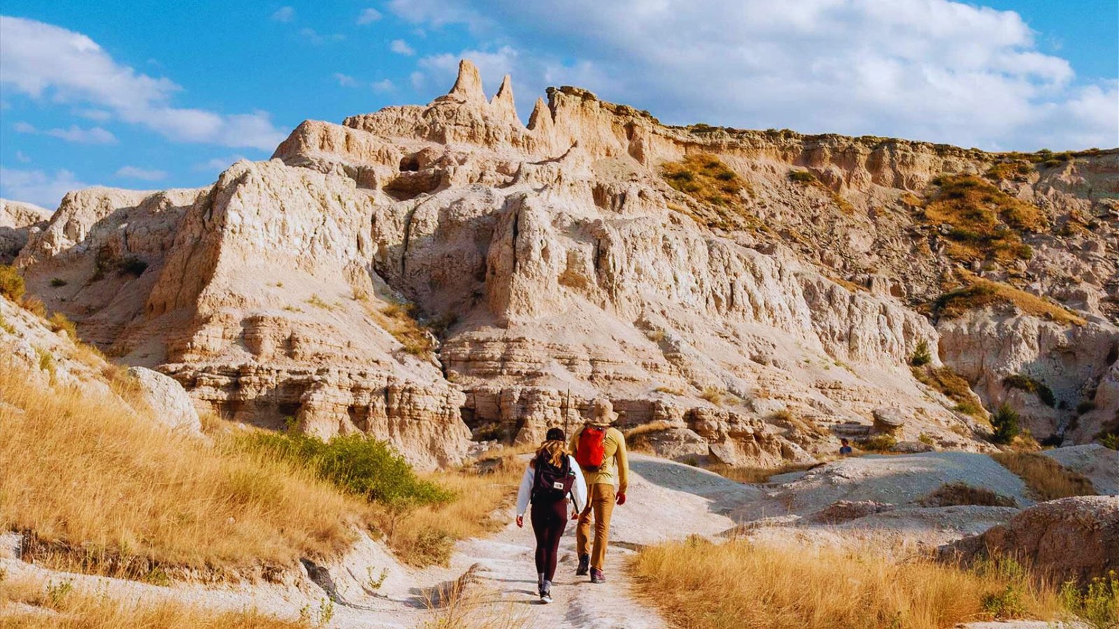 Two people walking along a sandy trail in a vast desert landscape under a clear blue sky.