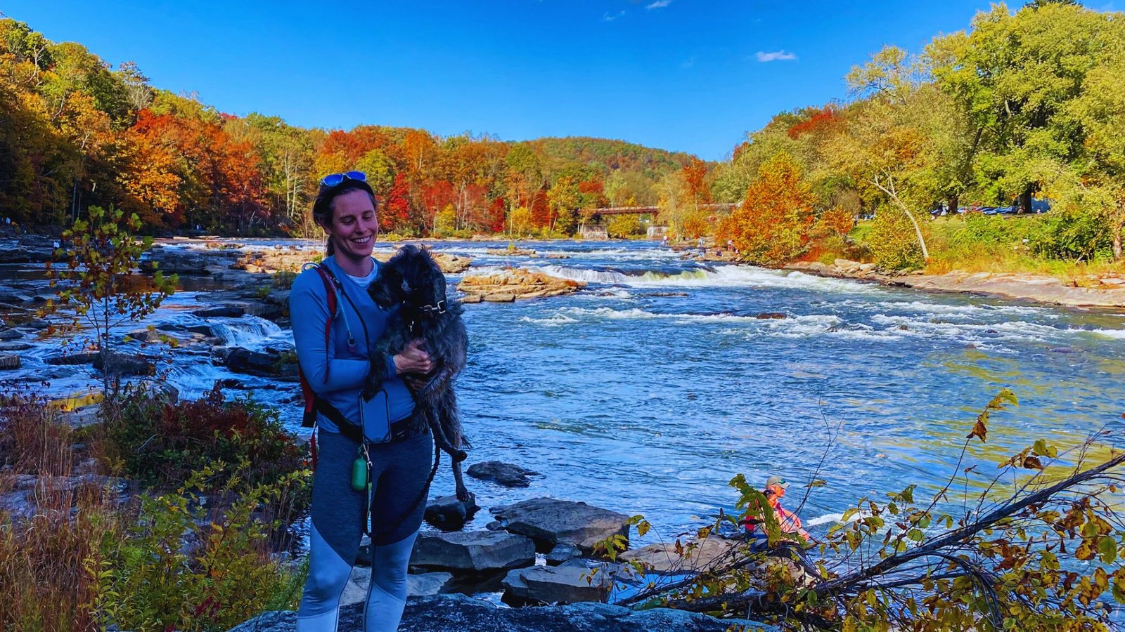  A woman stands beside a river with her dog, enjoying the serene outdoor setting.