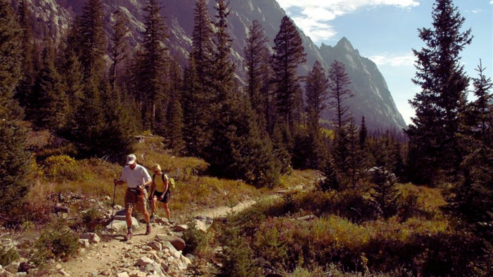 Two individuals hike along a scenic mountain trail, with towering peaks and vibrant foliage in the background.