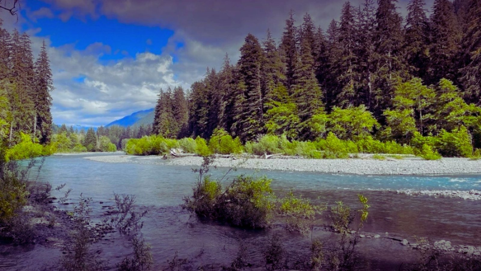 A serene river flows through a landscape of lush trees and majestic mountains in the background.