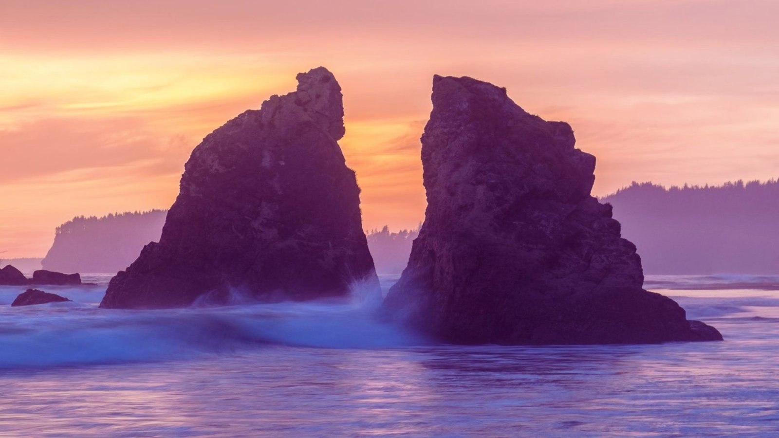 Two large rocks emerge from the ocean at sunset, casting silhouettes against a vibrant orange and purple sky.