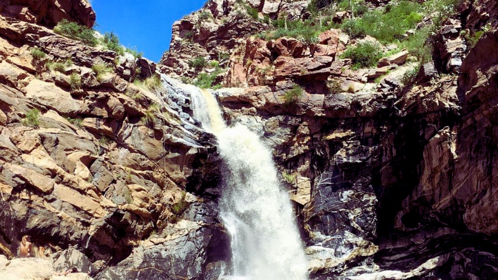 A waterfall cascades down a rugged mountain, surrounded by rocky terrain and lush greenery.
