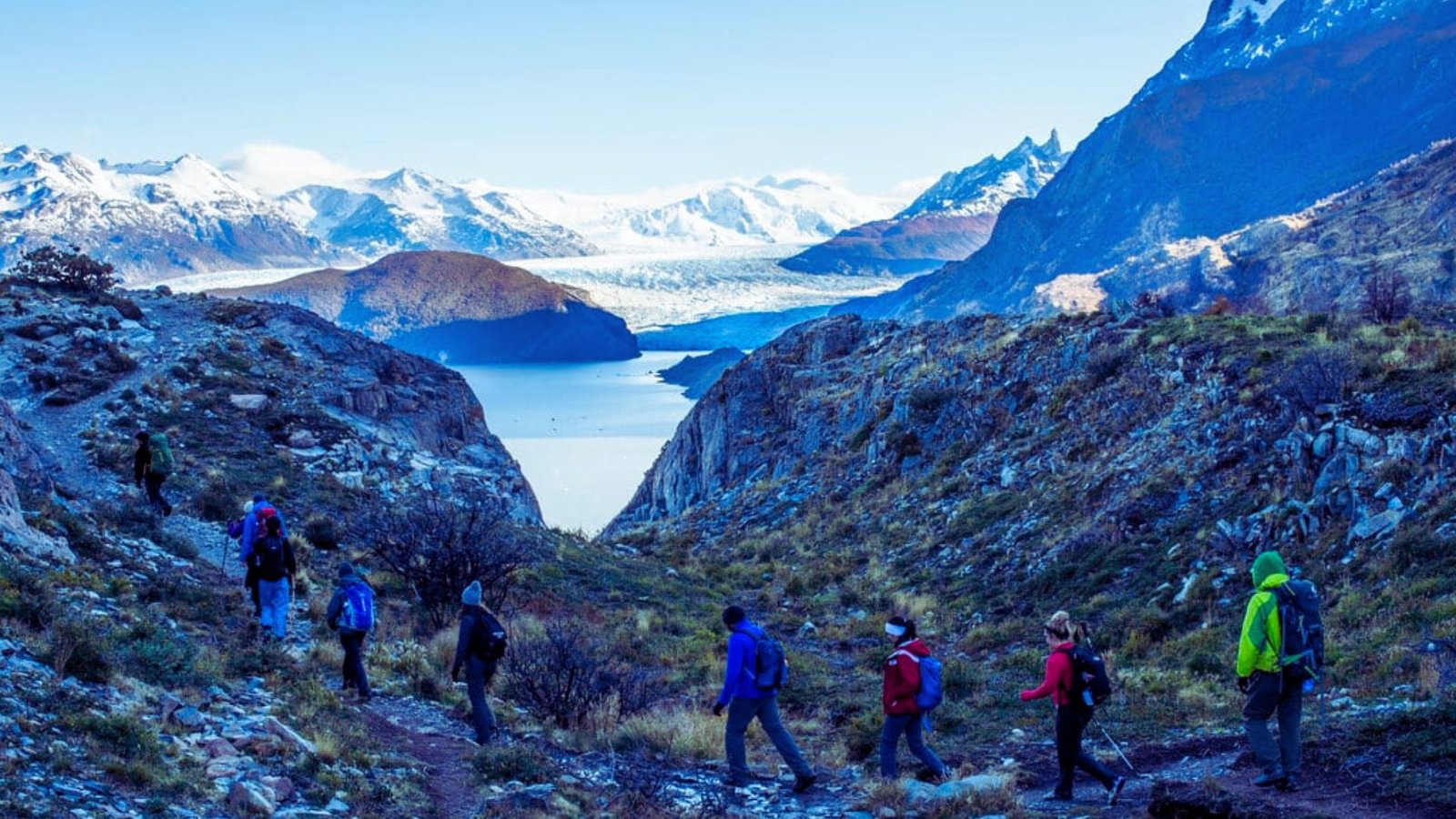  Hikers on a mountain trail with a serene lake and towering mountains in the background.
