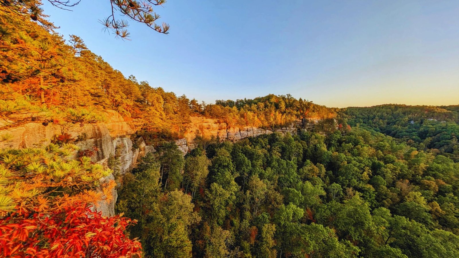 The sun sets behind rugged mountain cliffs, casting warm hues across the sky and illuminating the landscape.