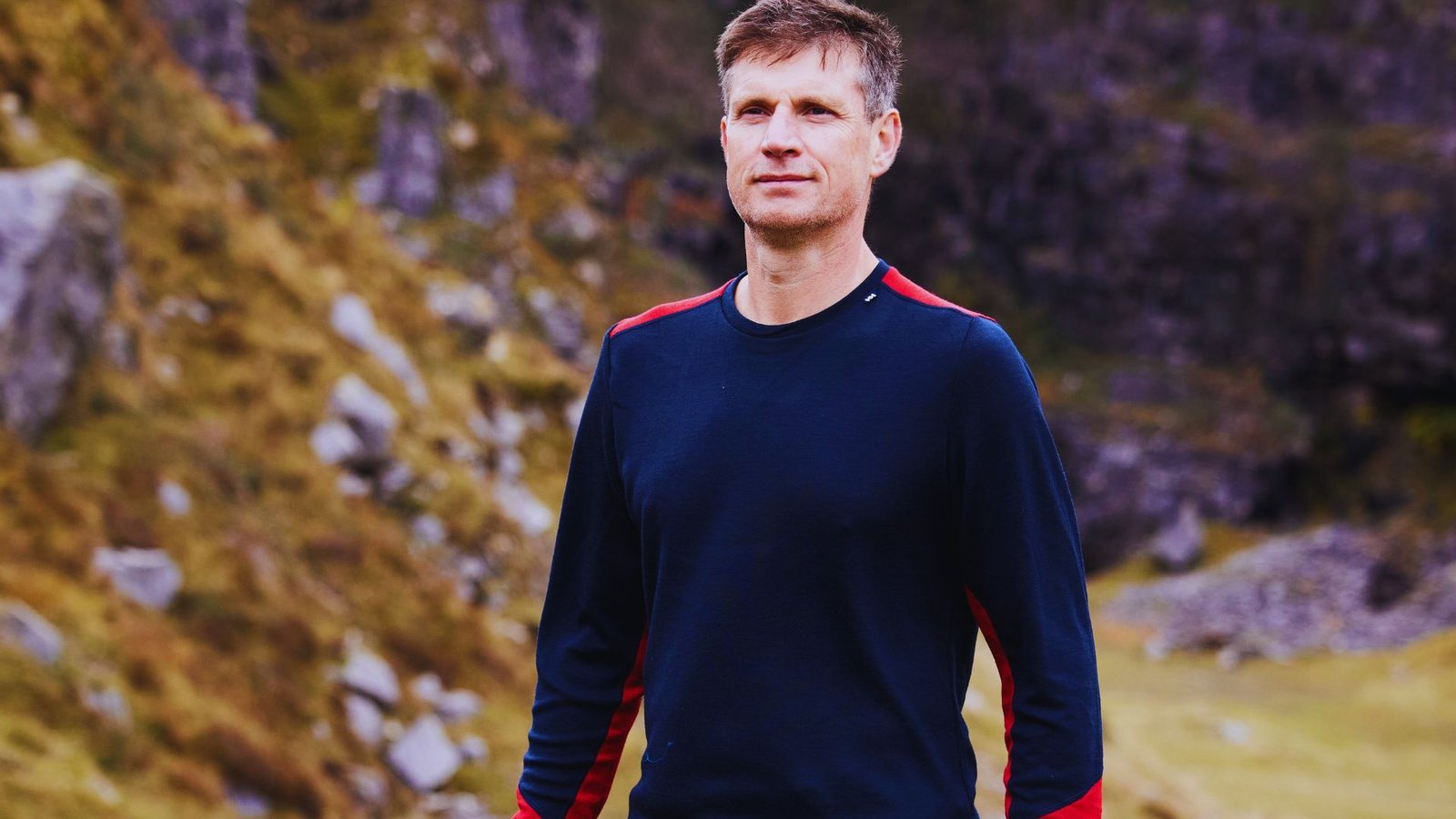 A man wearing a blue shirt and red pants stands on a rocky hill, gazing into the distance.