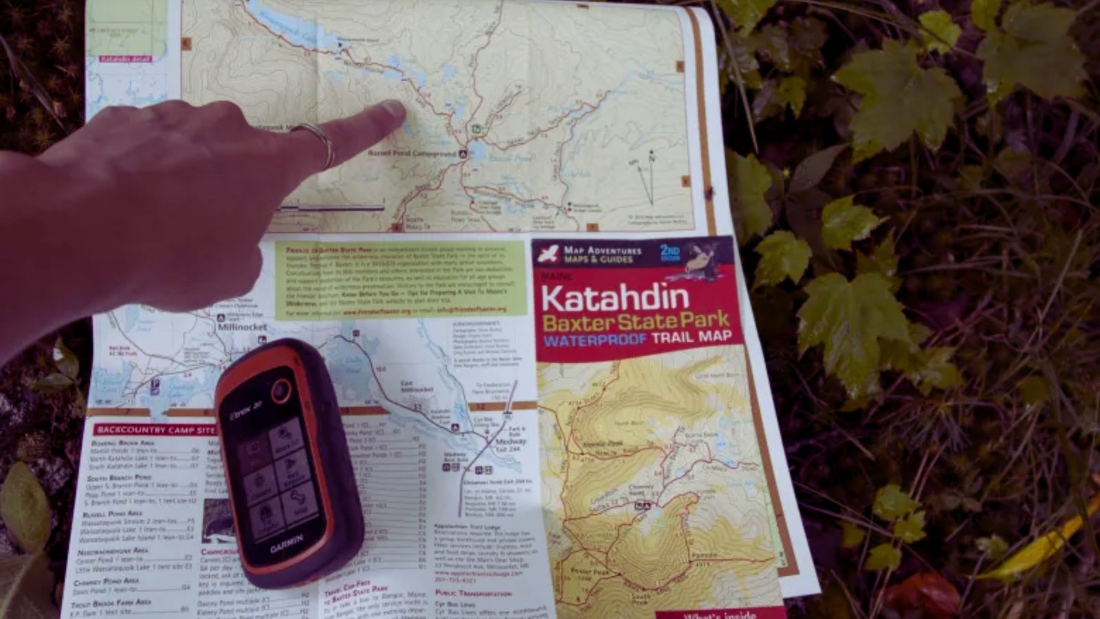 A person's hand gestures towards a map and GPS, suggesting directions or travel planning.