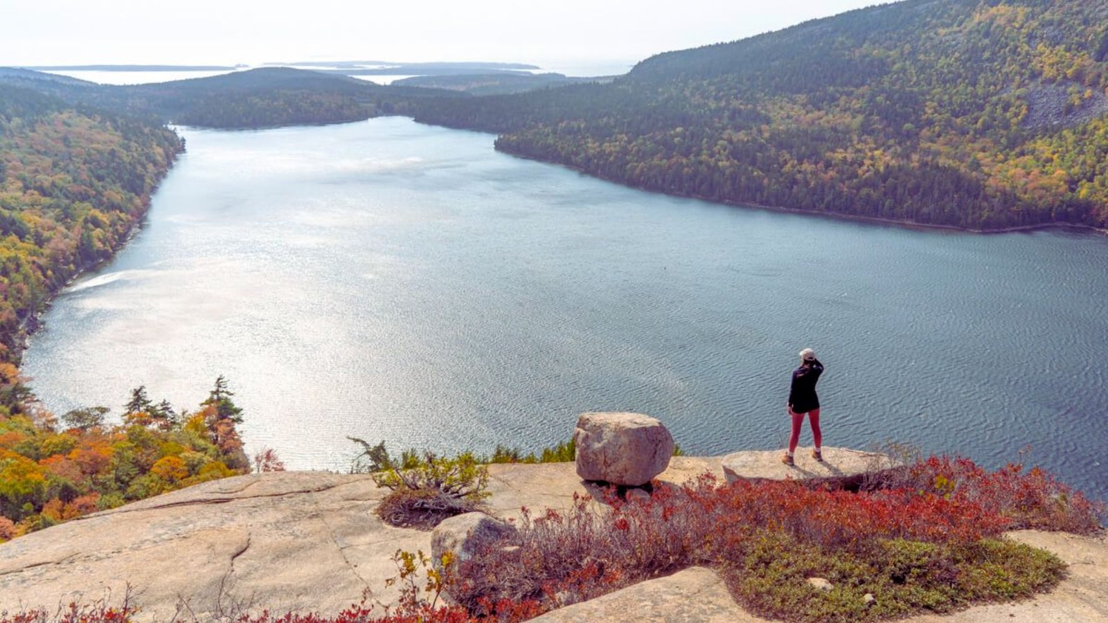 A person stands on a rock, gazing out over a serene lake surrounded by trees and mountains in the background.