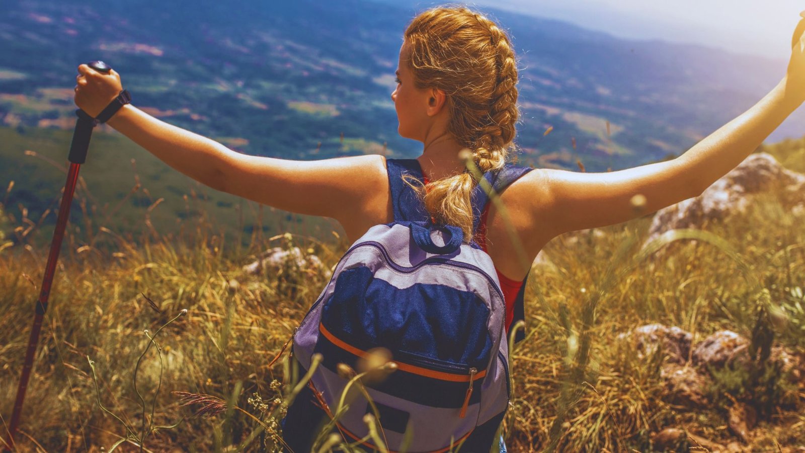  A woman with a backpack and hiking poles stands on a hill, arms outstretched, enjoying the view around her.
