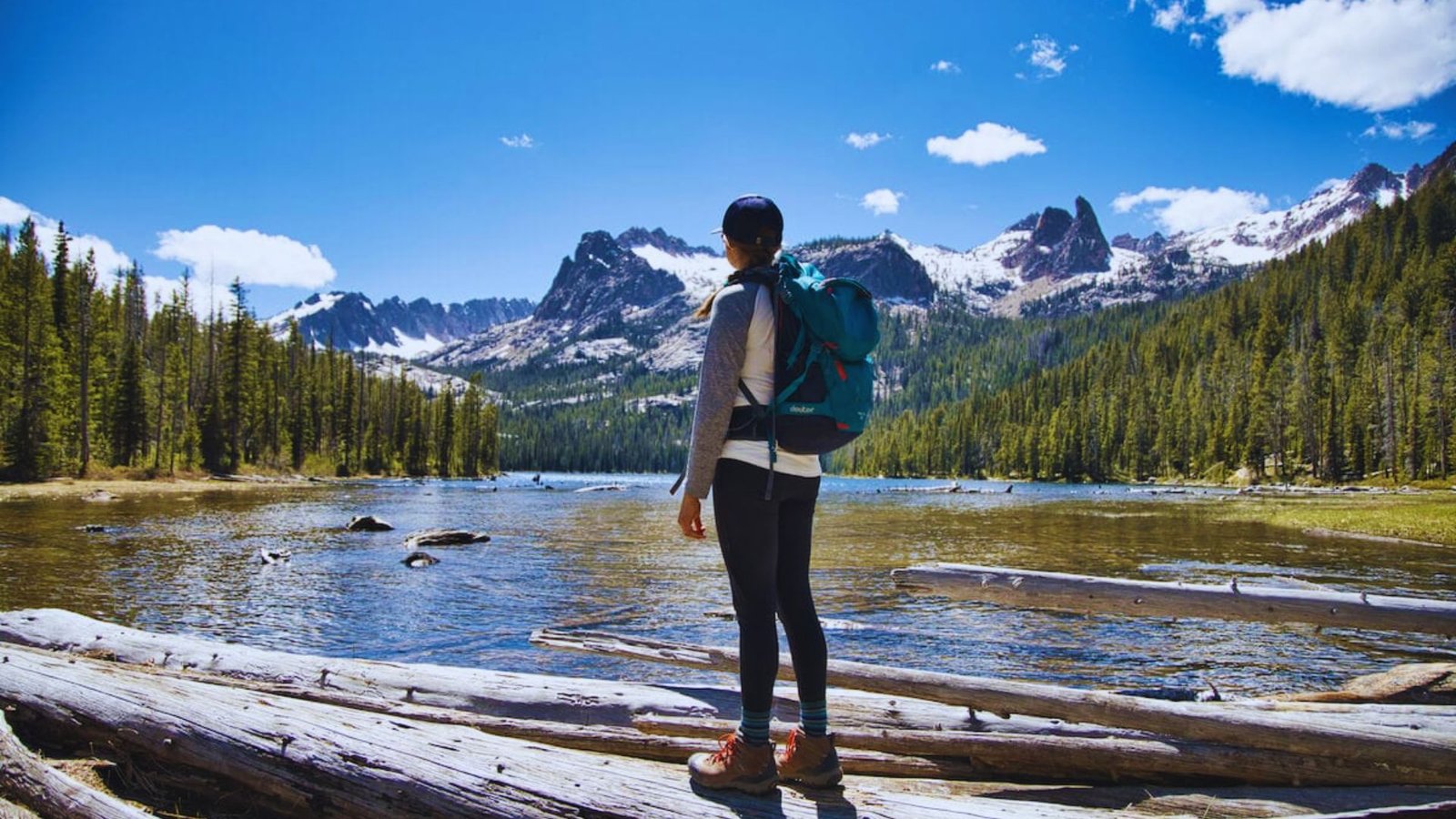 A woman with a backpack stands on a log, overlooking a serene lake surrounded by trees.