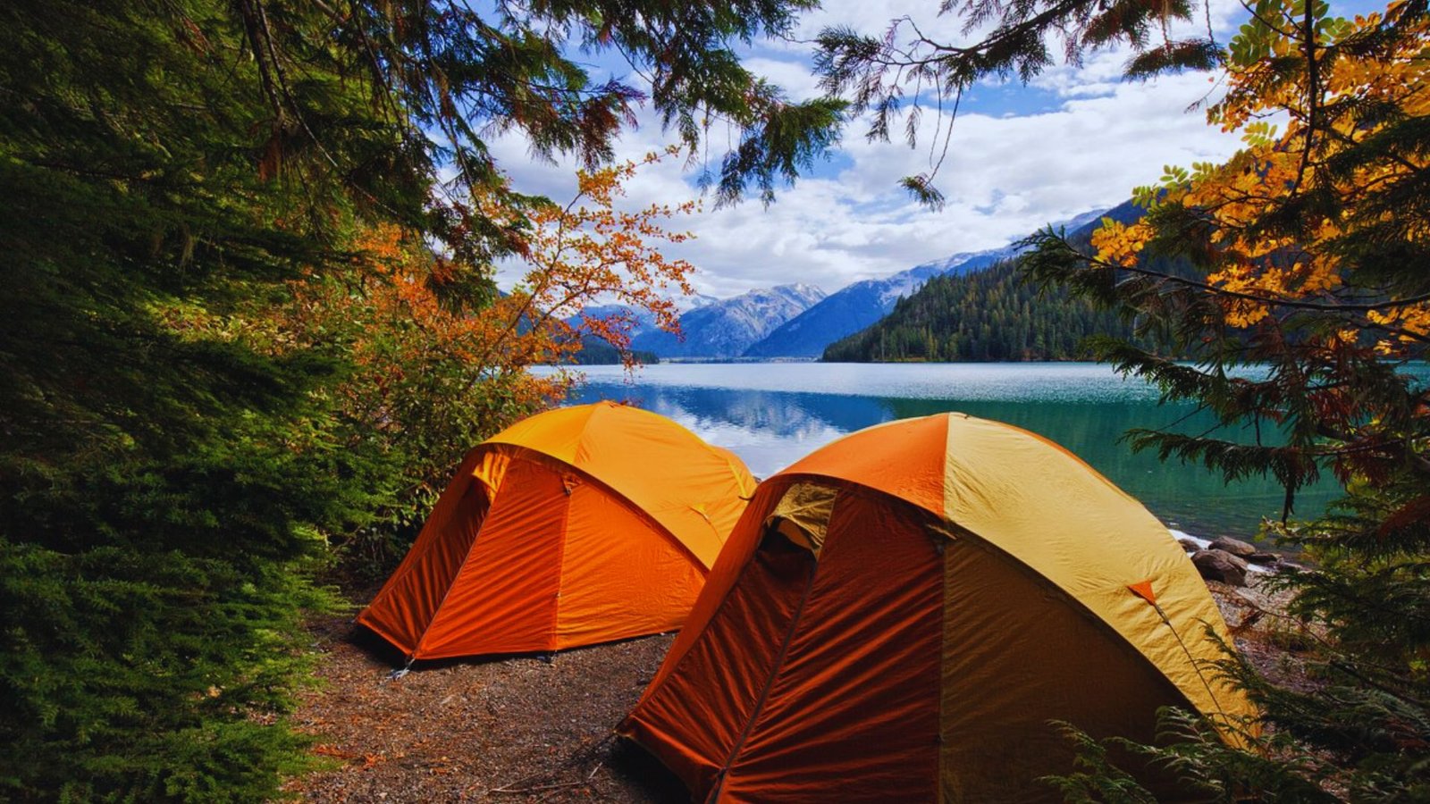 Two orange tents are pitched by a serene lake, with majestic mountains rising in the background, showcasing Michigan's beauty.