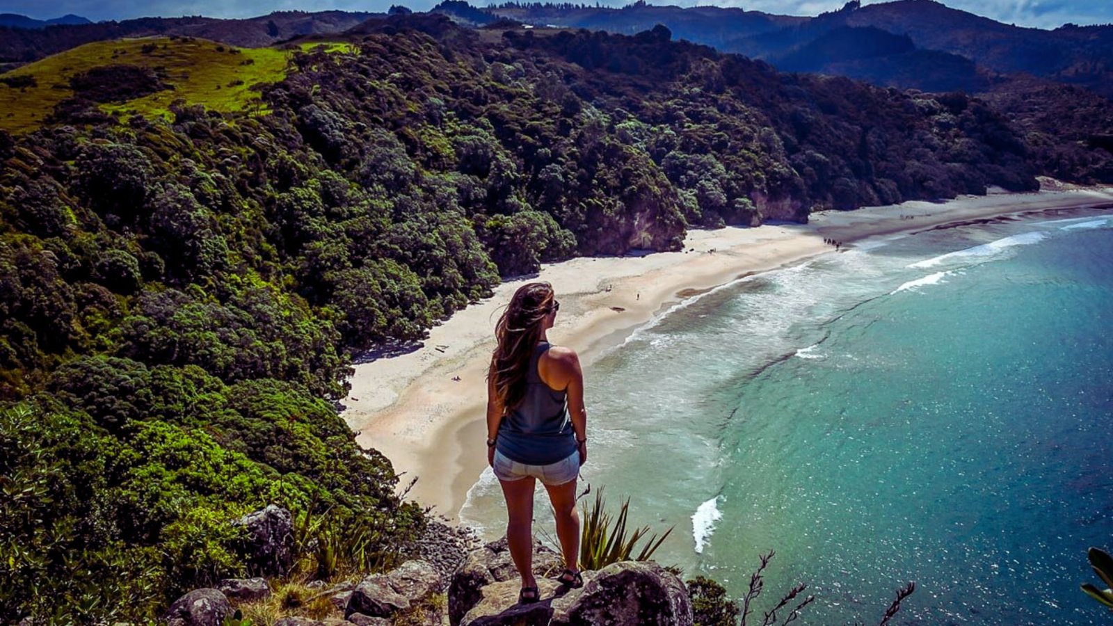 A woman stands on a cliff's edge, gazing at the beach below, with waves crashing against the shore in the distance.
