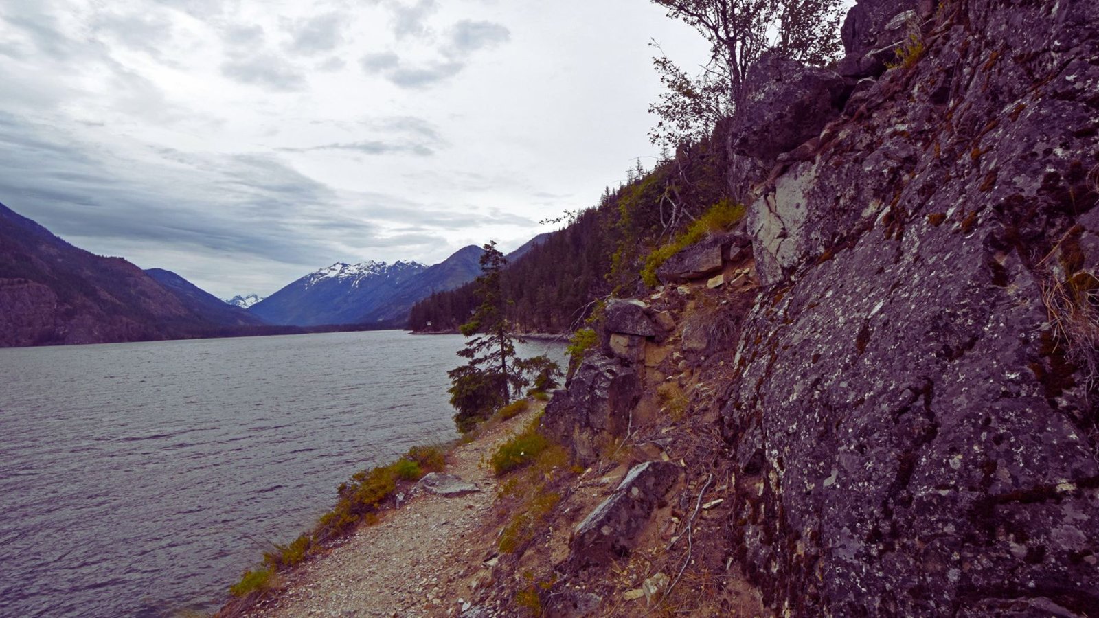  A rocky path winds toward a serene lake, framed by majestic mountains in the background.