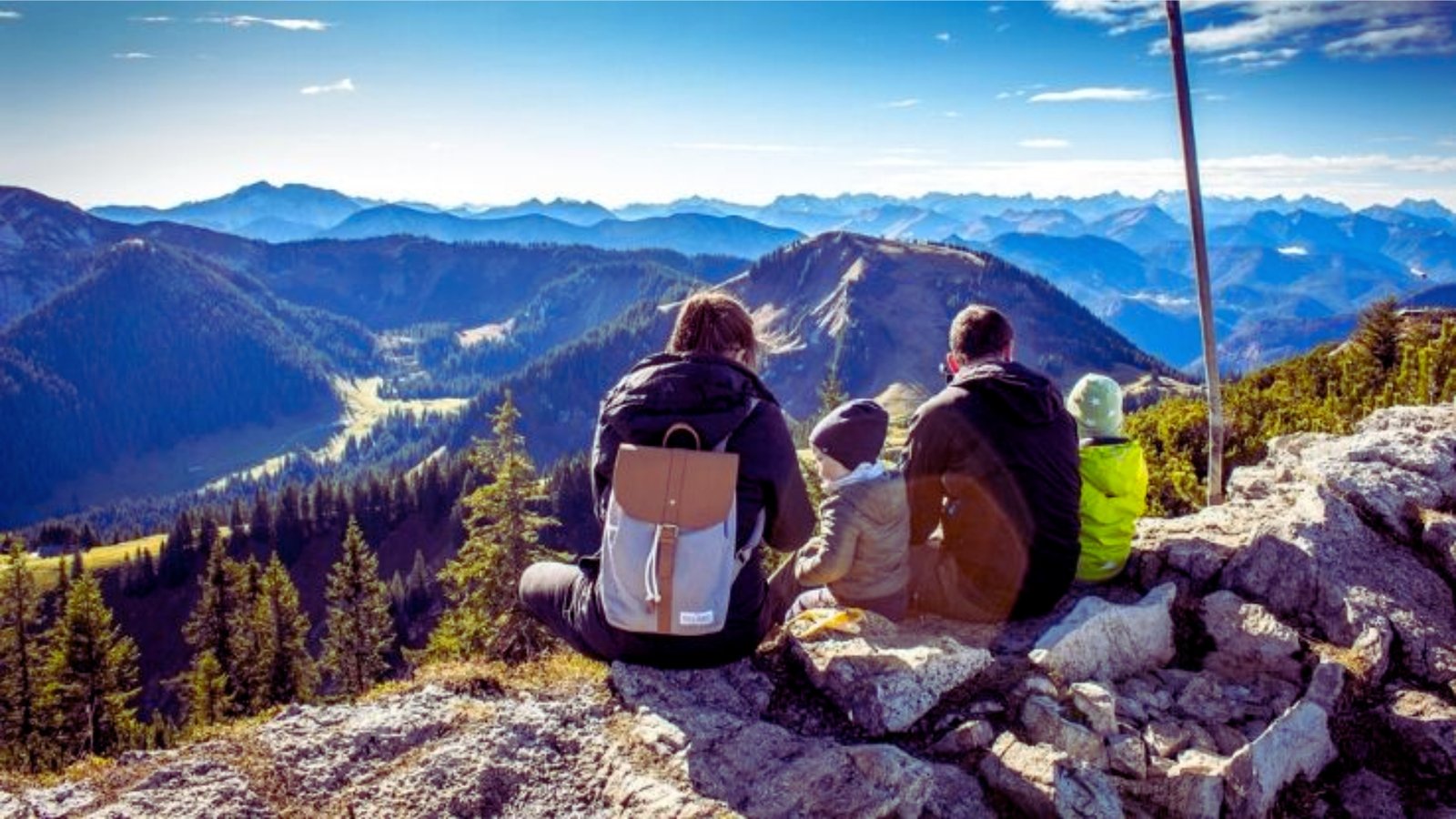 Family sitting on a mountain peak, gazing at the expansive view of valleys and distant hills.