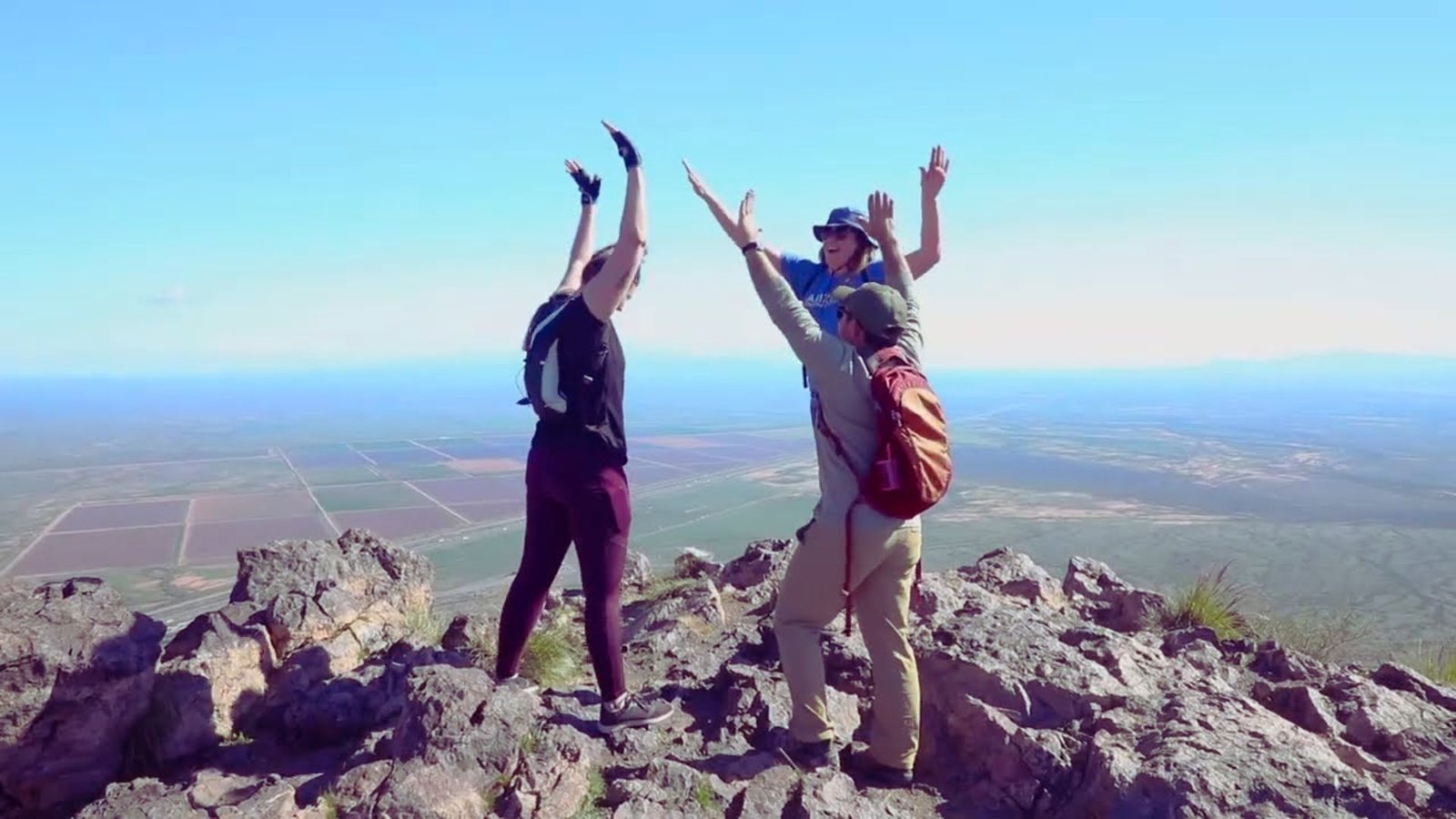 Three people stand triumphantly on a mountain peak, raising their hands in celebration against a clear blue sky.
