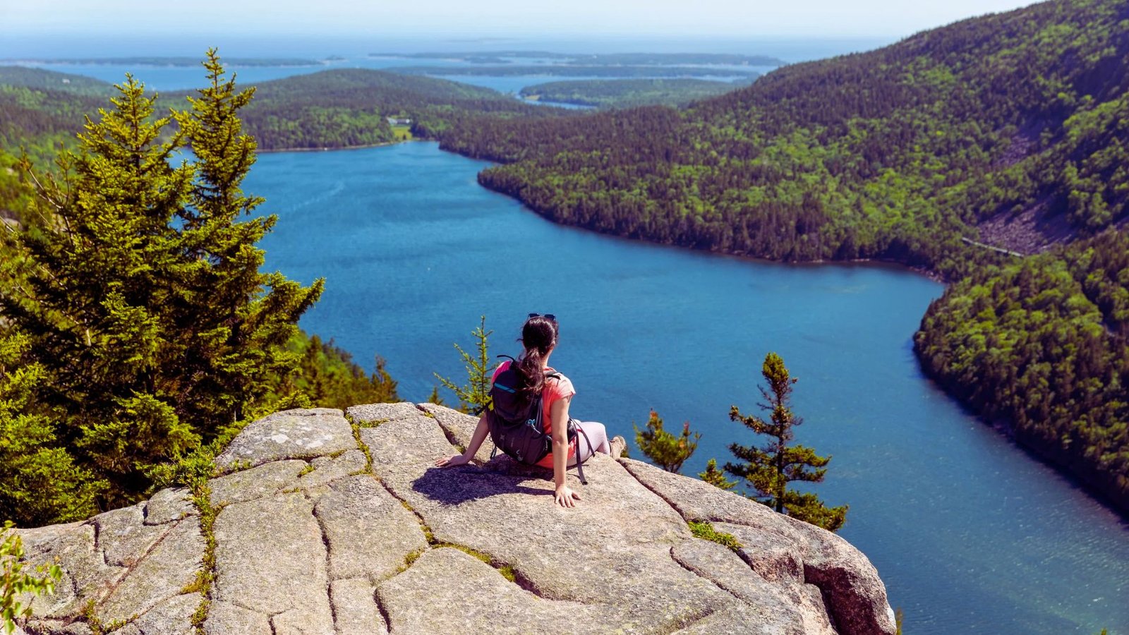 A person perched on a rock, overlooking a tranquil lake with lush greenery and distant hills in the background.