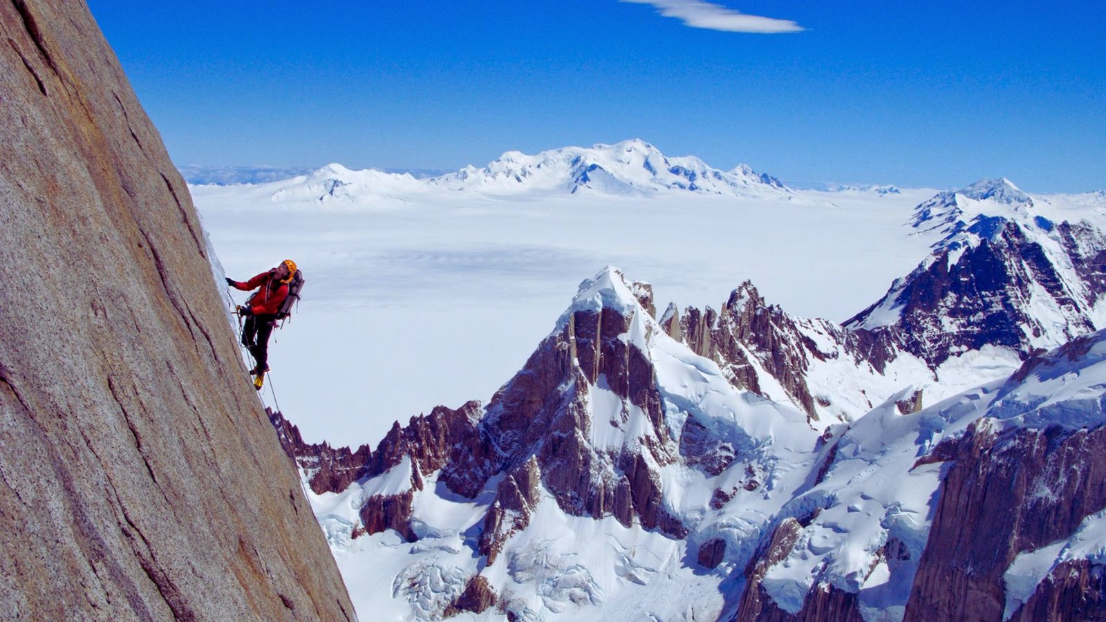 A climber ascends a rocky mountain, with snow-capped peaks visible in the background.
