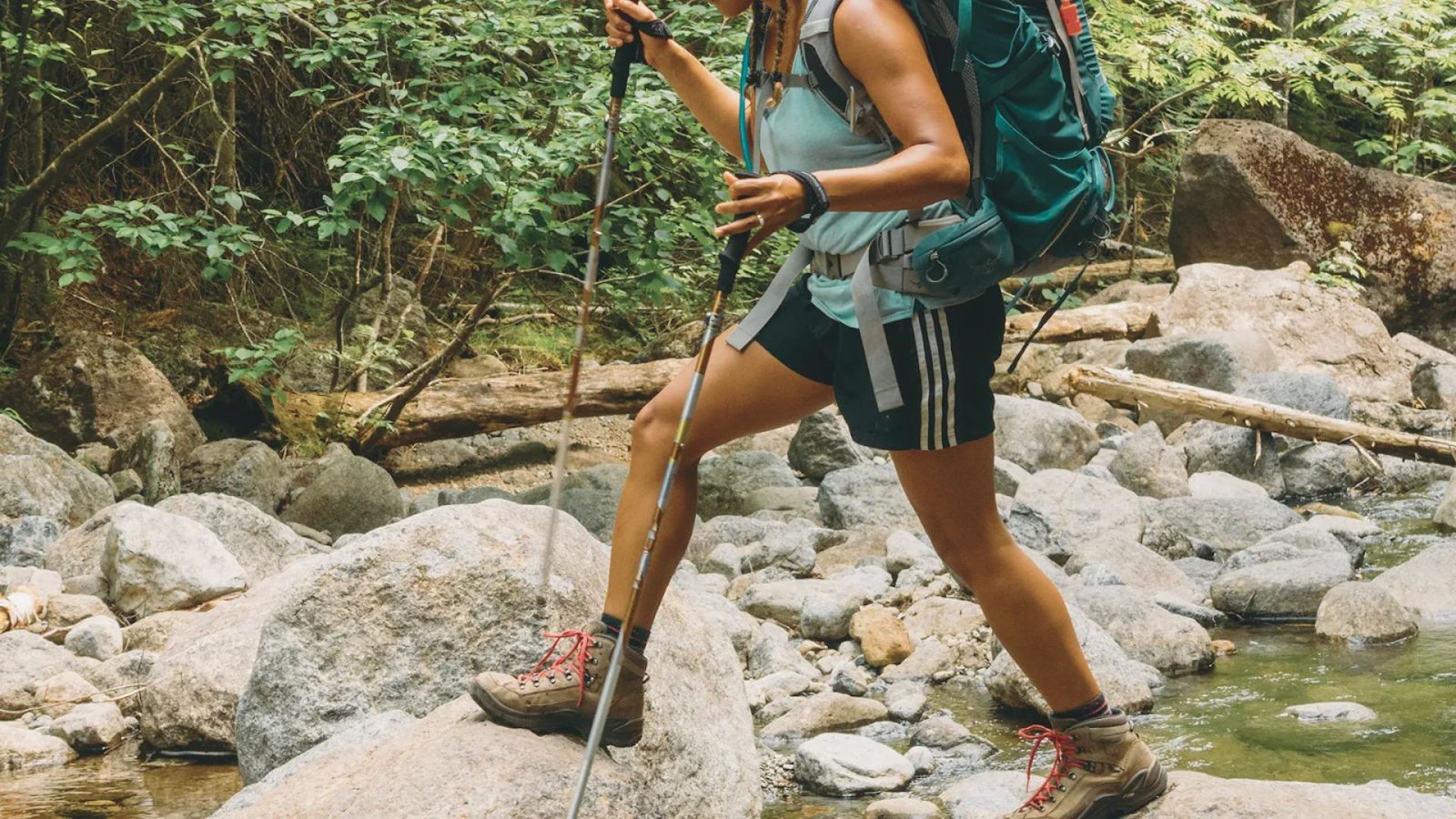 A woman with a backpack and hiking poles crosses a stream, navigating the rocky terrain with care.