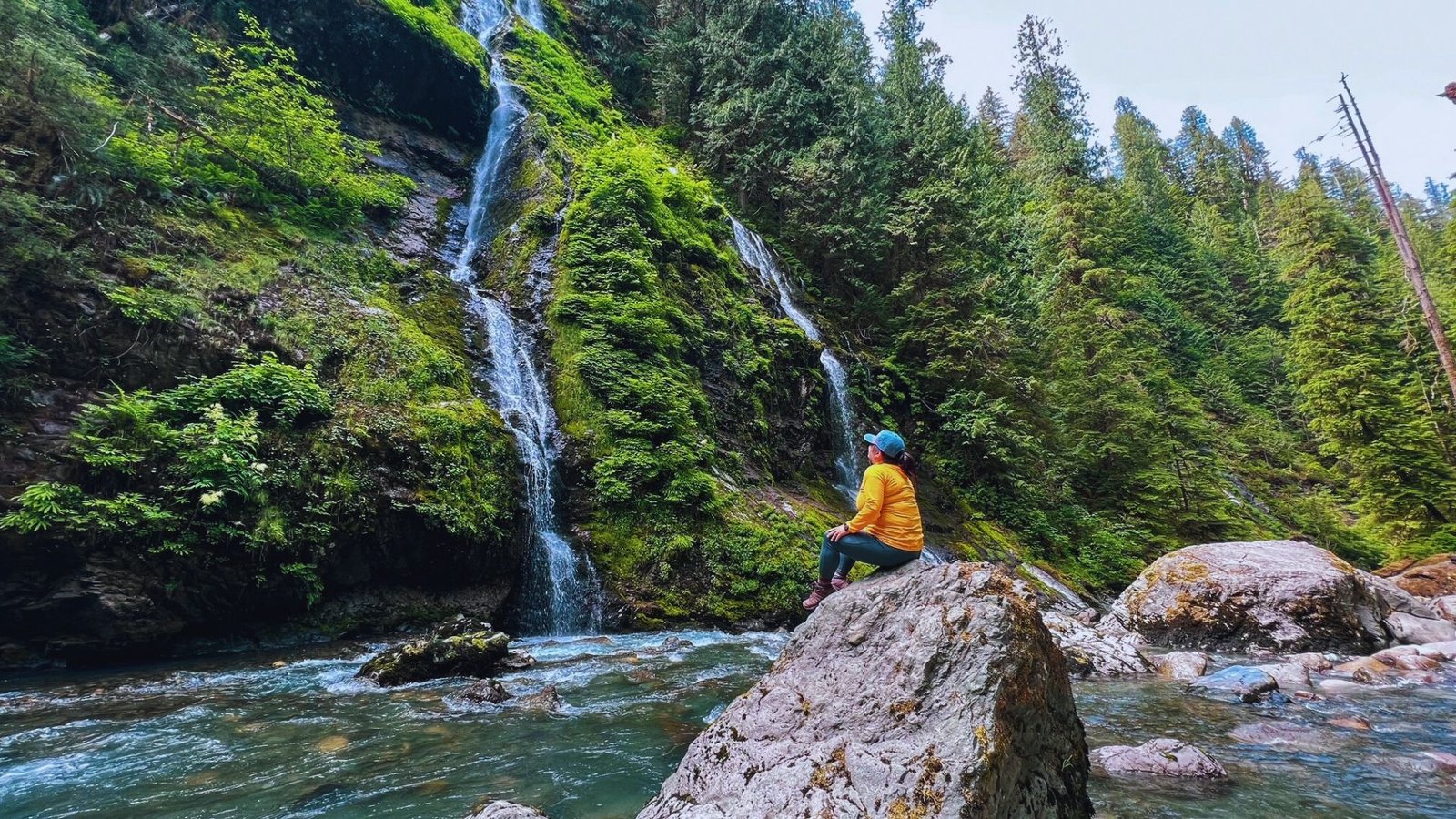  A person sits on a rock beside a flowing waterfall along the Boulder River Trail, surrounded by lush greenery.
