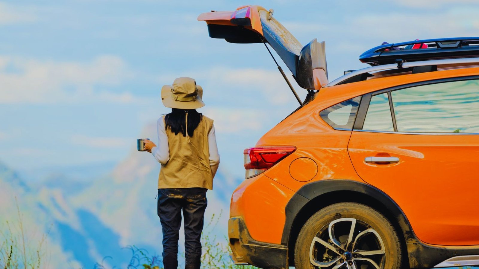 A woman stands beside an orange convertible car, ready for a road trip, with the top down and a scenic background.